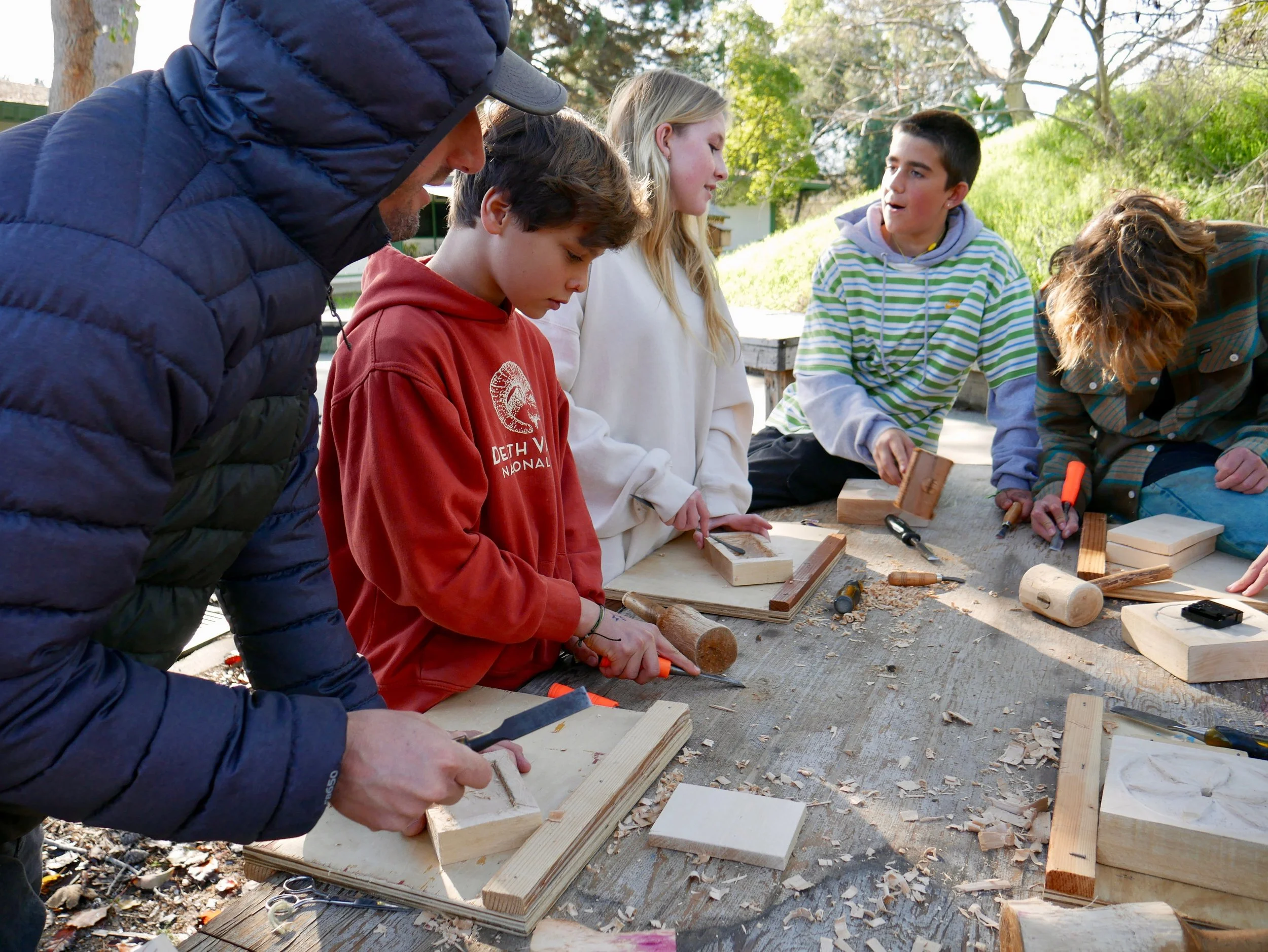 A group of six people working on woodworking projects outdoors on a sunny day, using hand tools on wooden pieces.