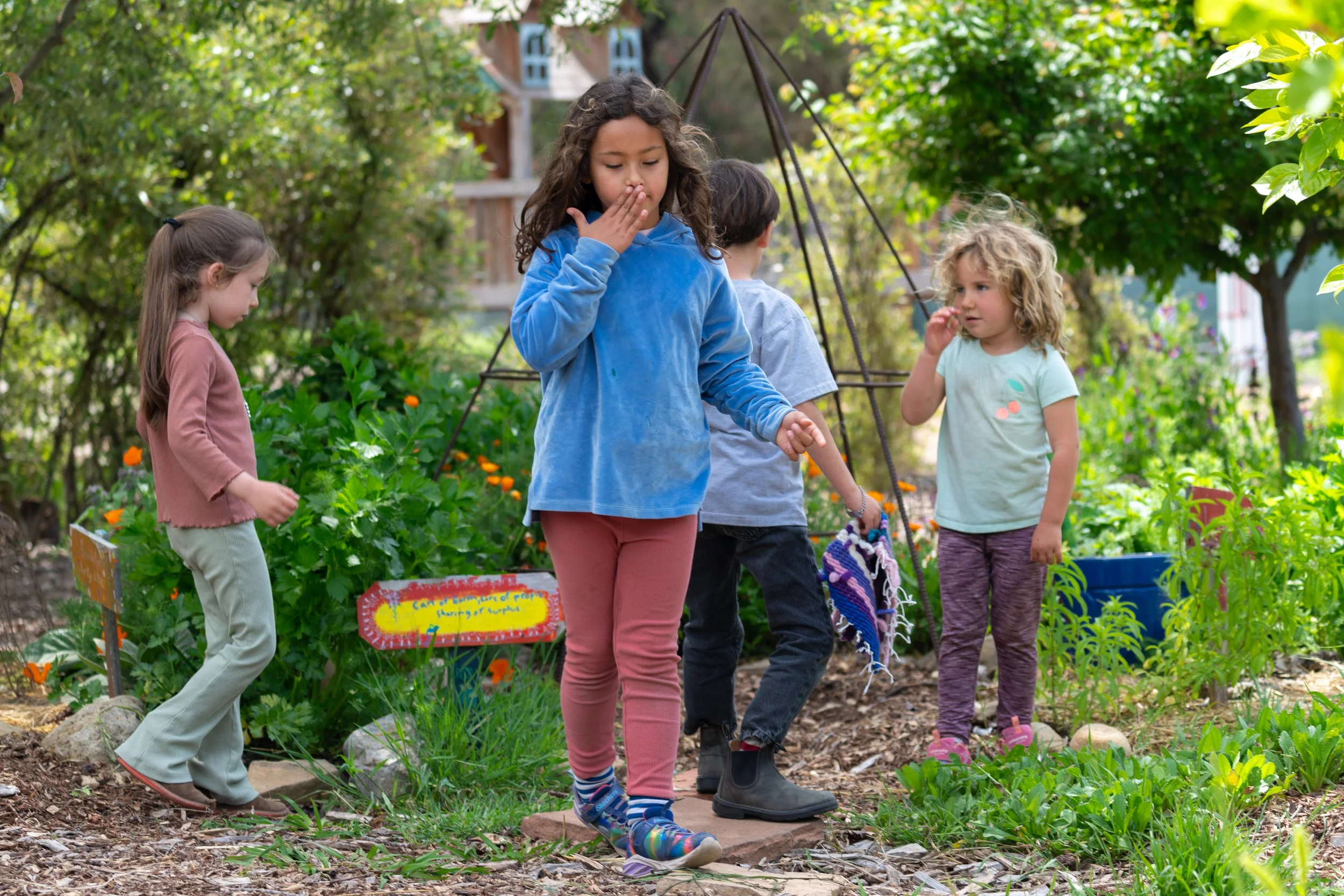 Four children exploring a garden with lush green plants and flowers, some signs and rain boots on a stone path.