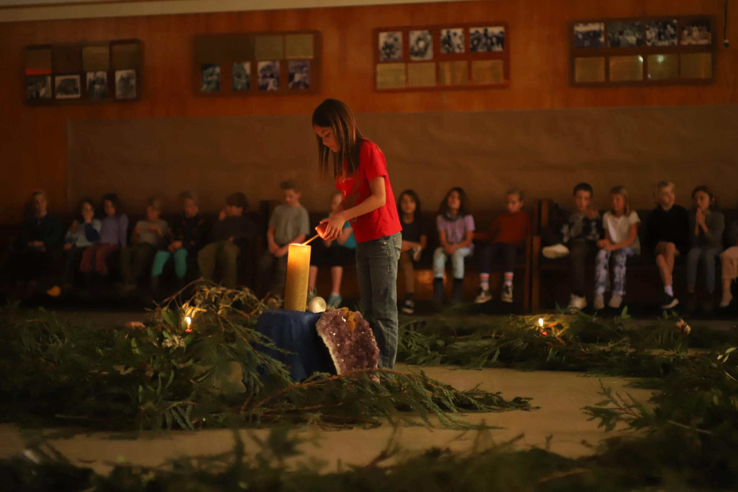 A girl lighting a candle in a dimly lit hall with children sitting on benches, decorated with greenery and illuminated objects, possibly during a ceremony or event.