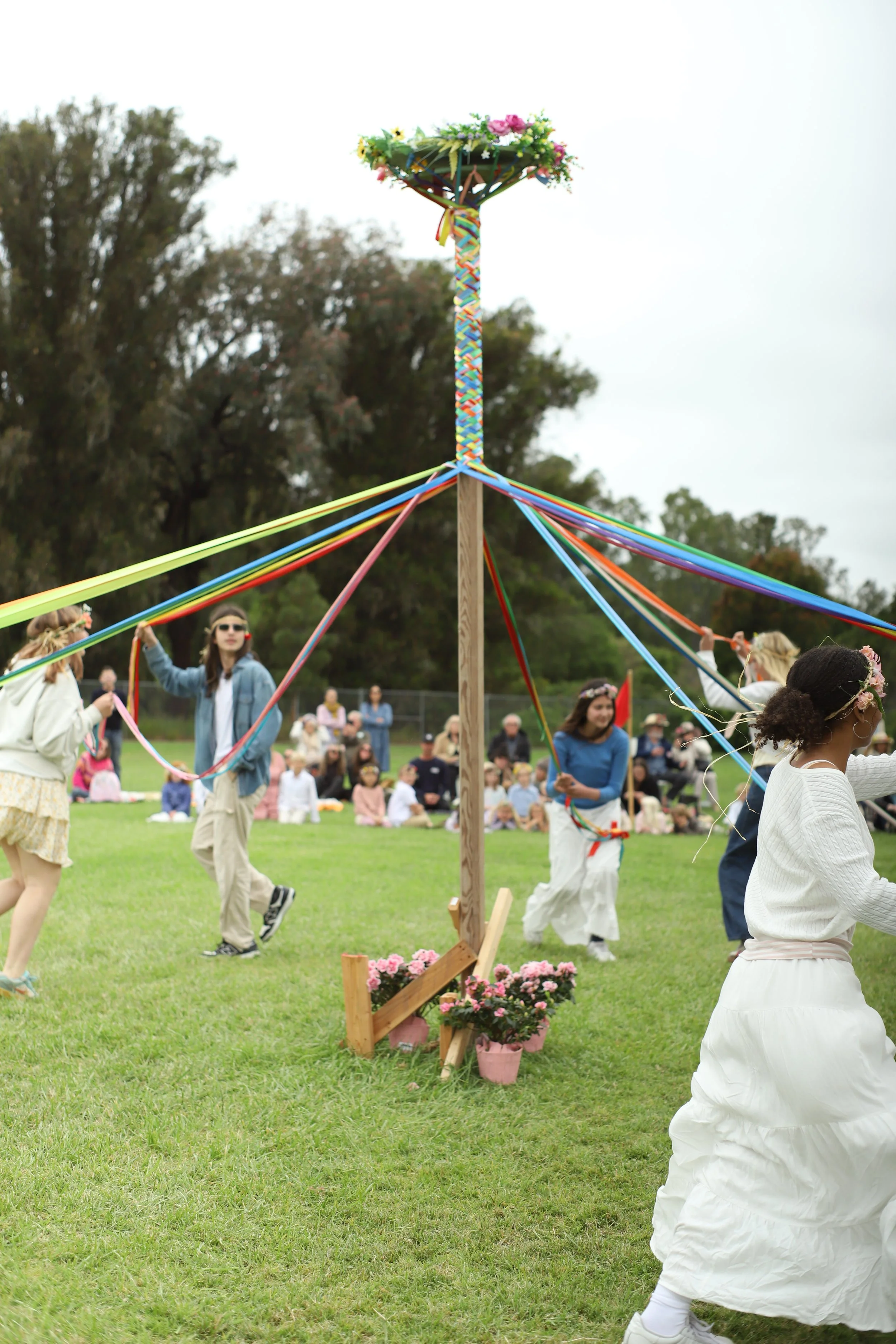 People dancing around a maypole decorated with colorful ribbons and flowers during a celebration in an open grassy park, with an audience seated in the background.