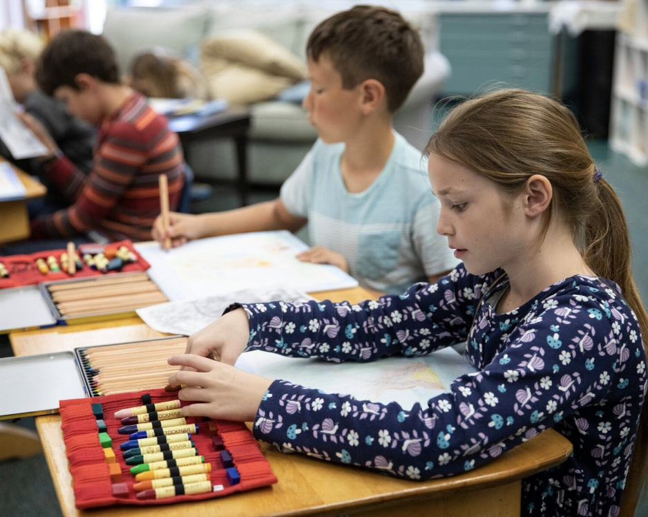 A classroom scene with children working on a drawing activity, using colored pencils on a wooden table.