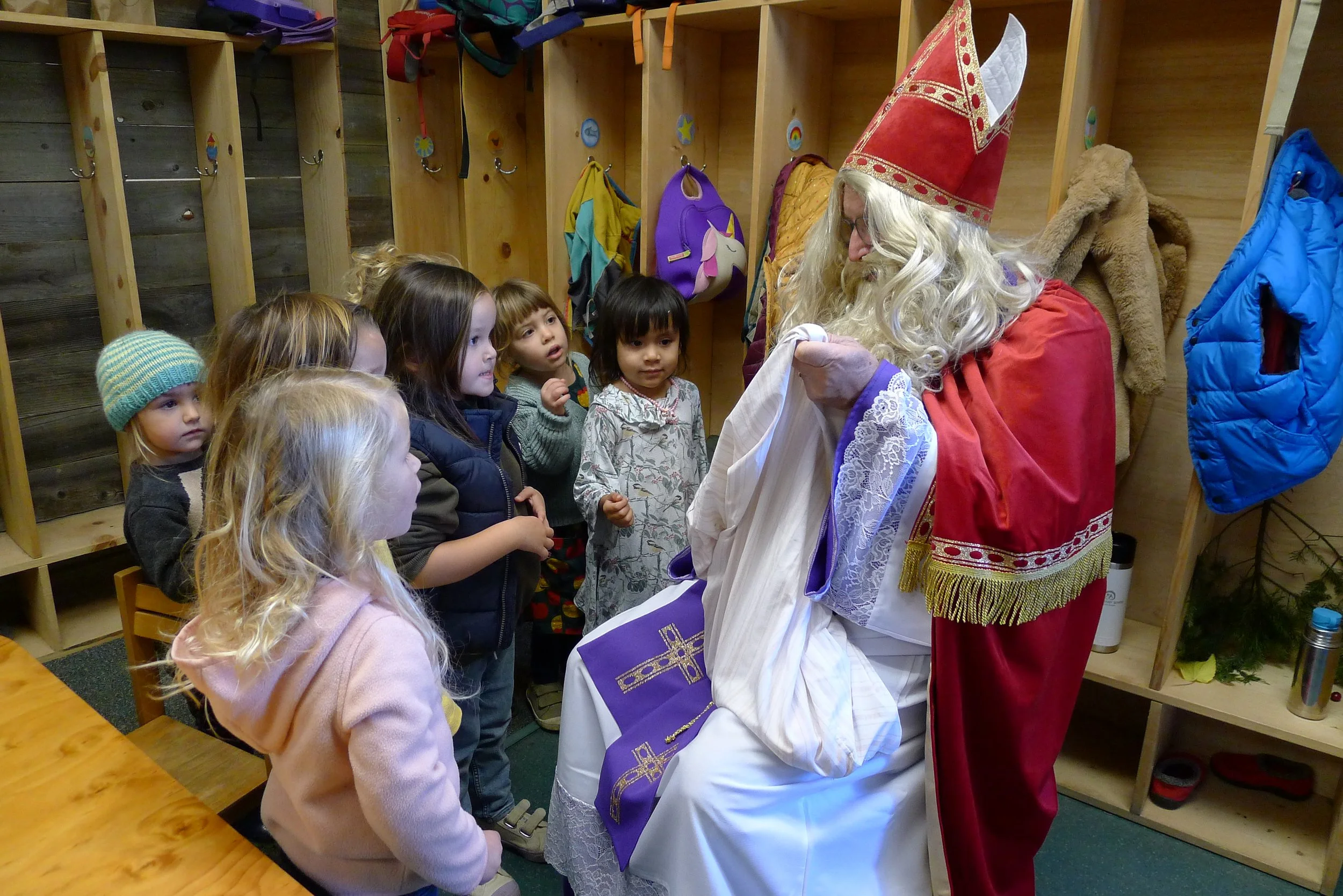 Children participating in a classroom holiday celebration, with some dressed in white robes and hats resembling a Christmas pageant, carrying candles and a gift, while others observe at their desks.