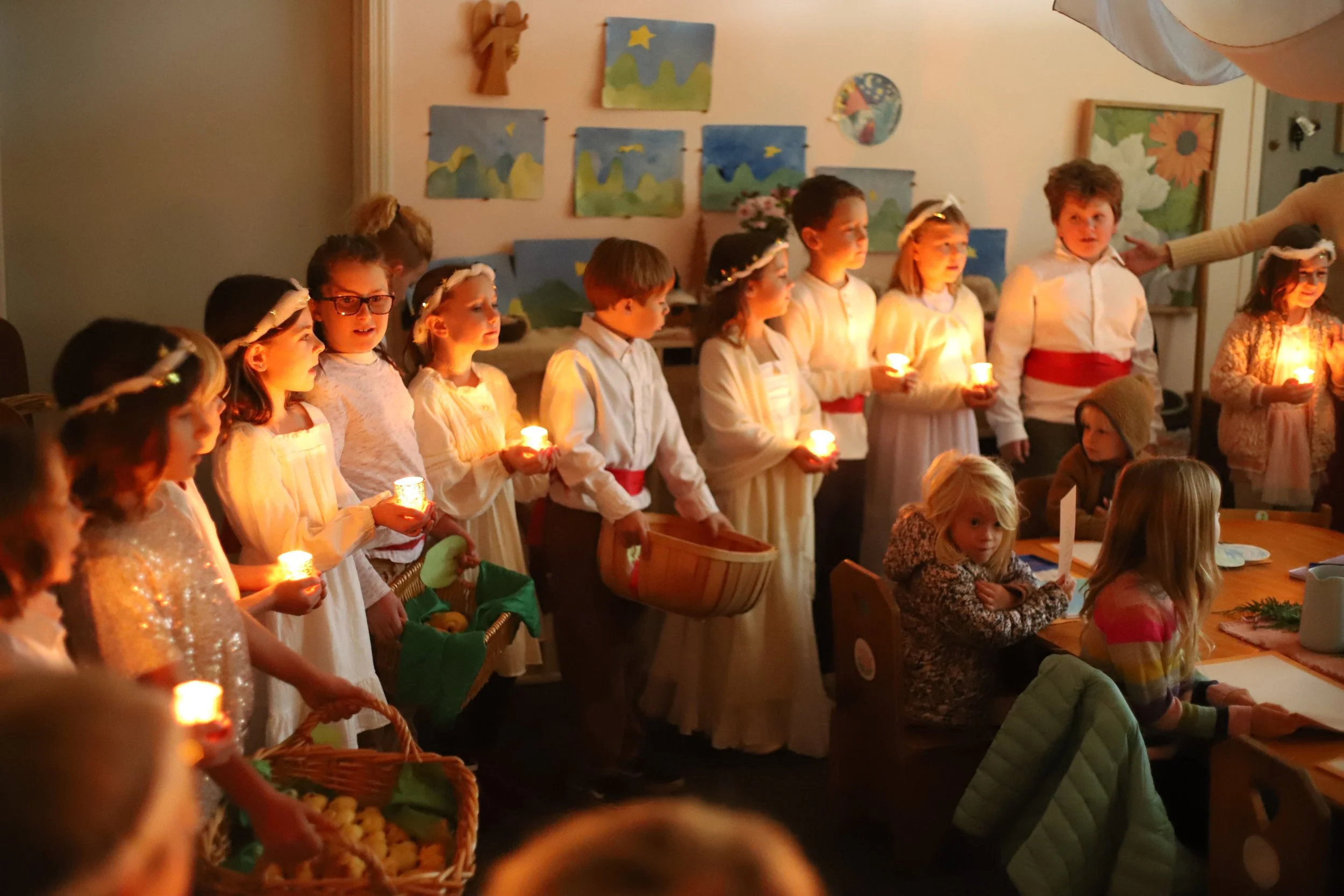 Children dressed in white robes, wearing headbands with halos, holding candles, standing in a line during a Christmas pageant or celebration, with a woman handing out a candle, in a decorated room with children's artwork on the walls.