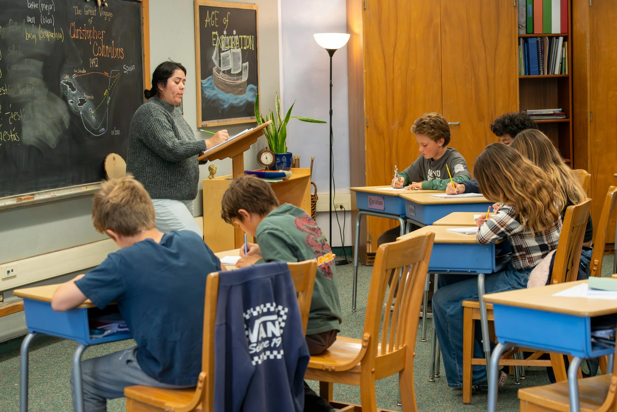 A classroom with a teacher standing at a podium writing in a notebook, while students sit at desks taking notes. The blackboard has drawings and text about Christopher Columbus and exploration.