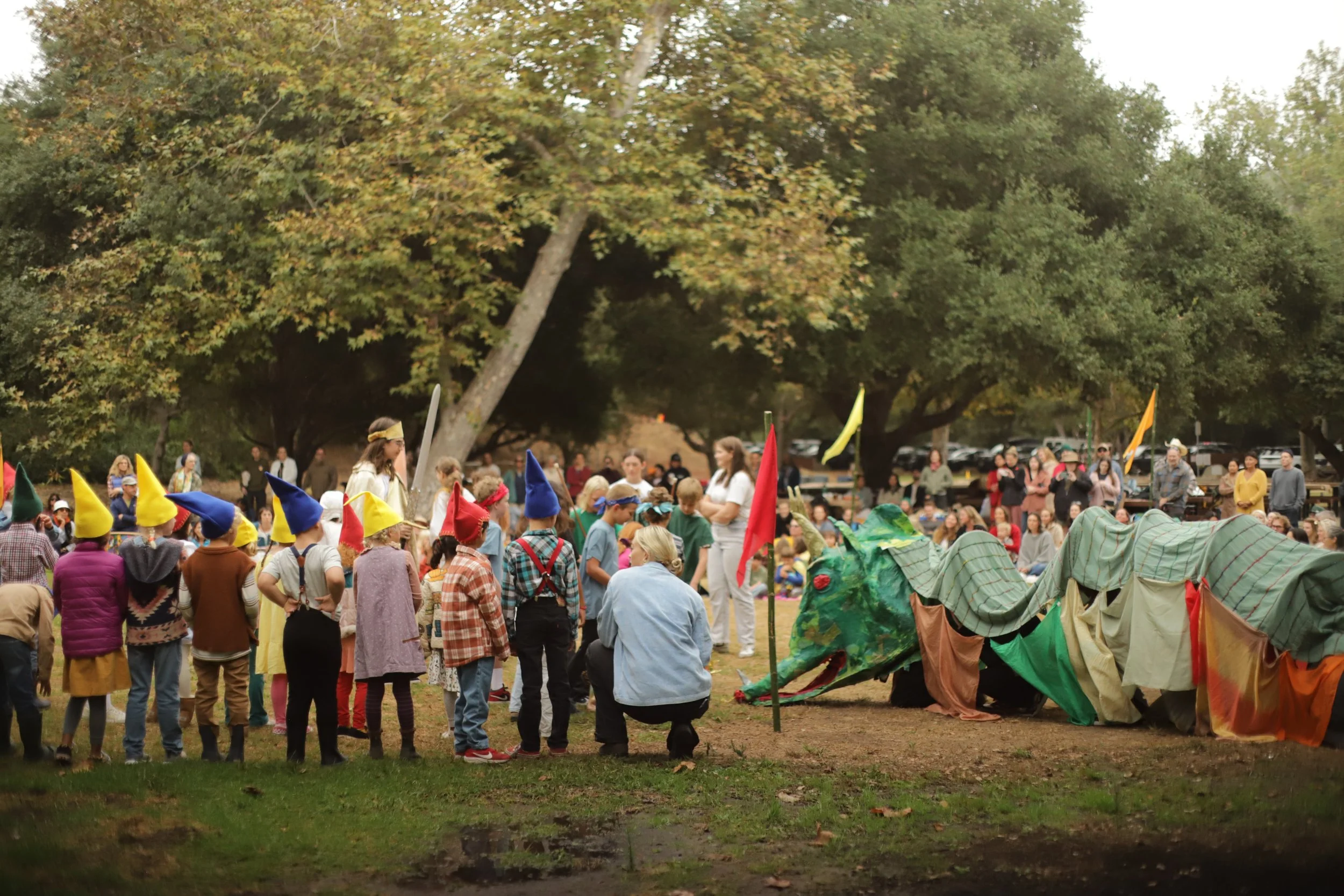 People gathered outdoors for a puppet show featuring a large colorful dragon, with participants wearing whimsical hats, including a row of children with yellow, blue, and red pointy hats, in a park with trees and onlookers in the background.