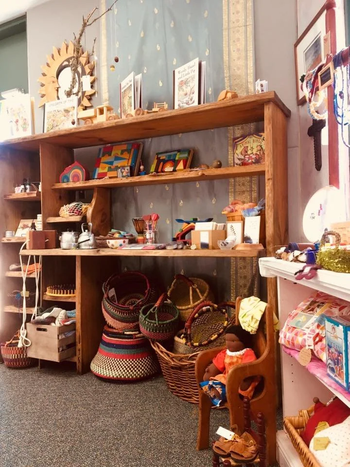Display of handmade crafts on wooden shelves and baskets, including jewelry, dolls, woven baskets, and decorative items, in a cozy indoor shop.