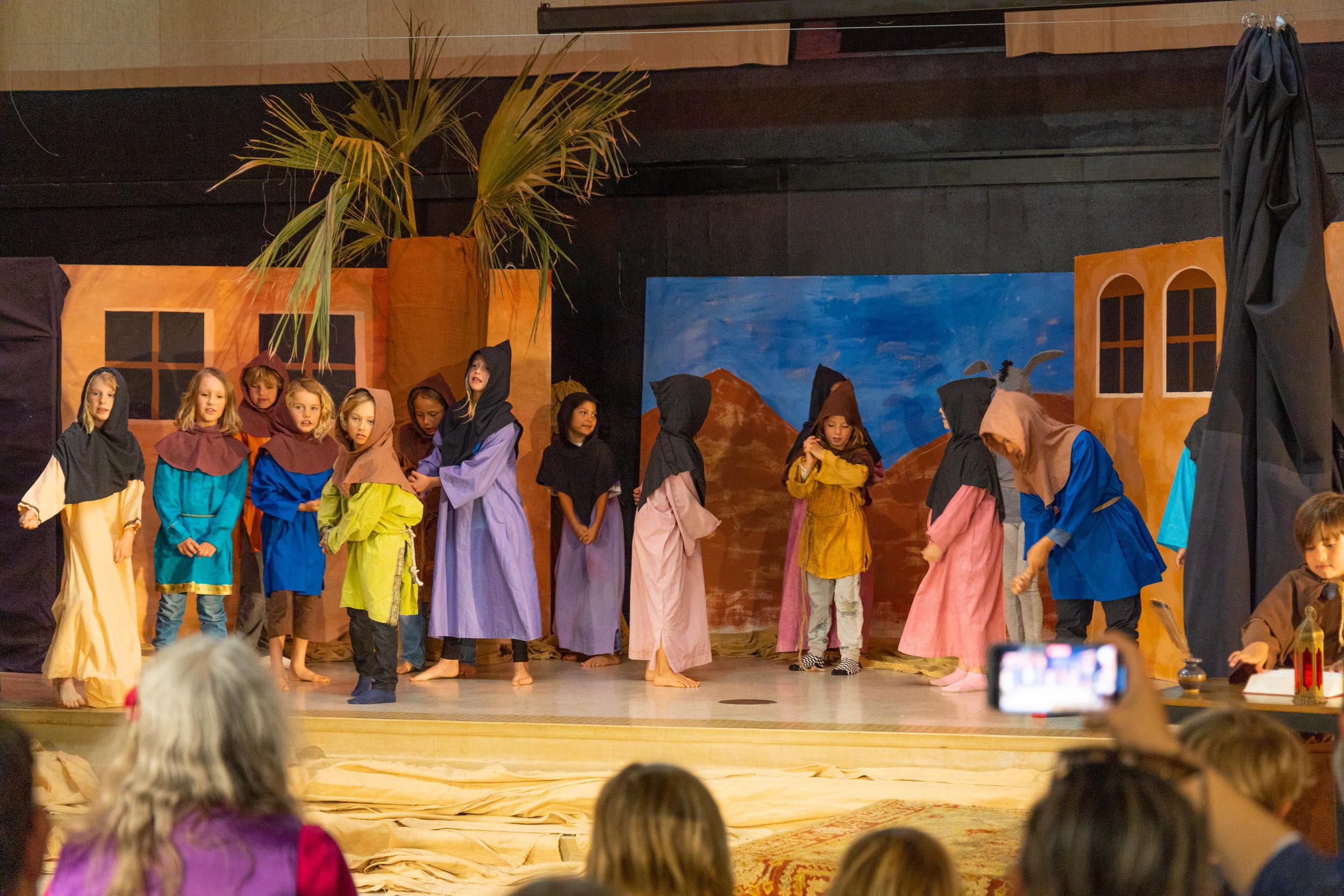 Children performing a play on stage, dressed in costumes resembling biblical or historical characters, with a painted backdrop of mountains, a blue sky, and windows, while an audience watches.
