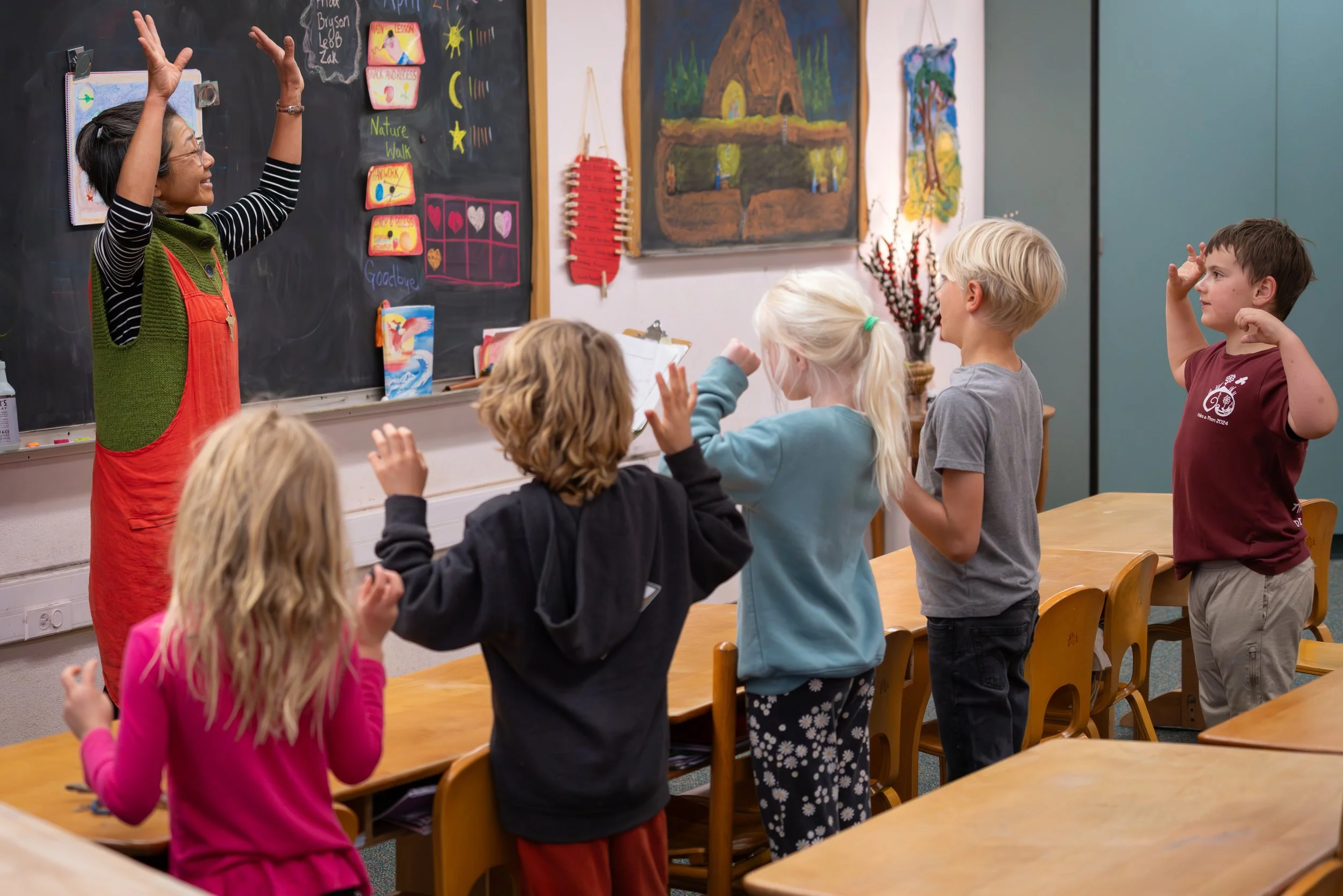 A teacher in a classroom standing in front of a blackboard, interacting with young students who are raising their hands. The classroom has colorful artwork and educational posters on the walls.