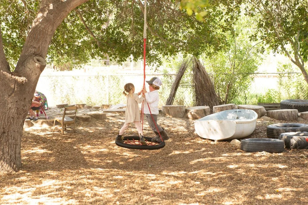 Two children, a girl and a boy, playing on a tire swing in a backyard with trees, chairs, a bathtub, and tires.