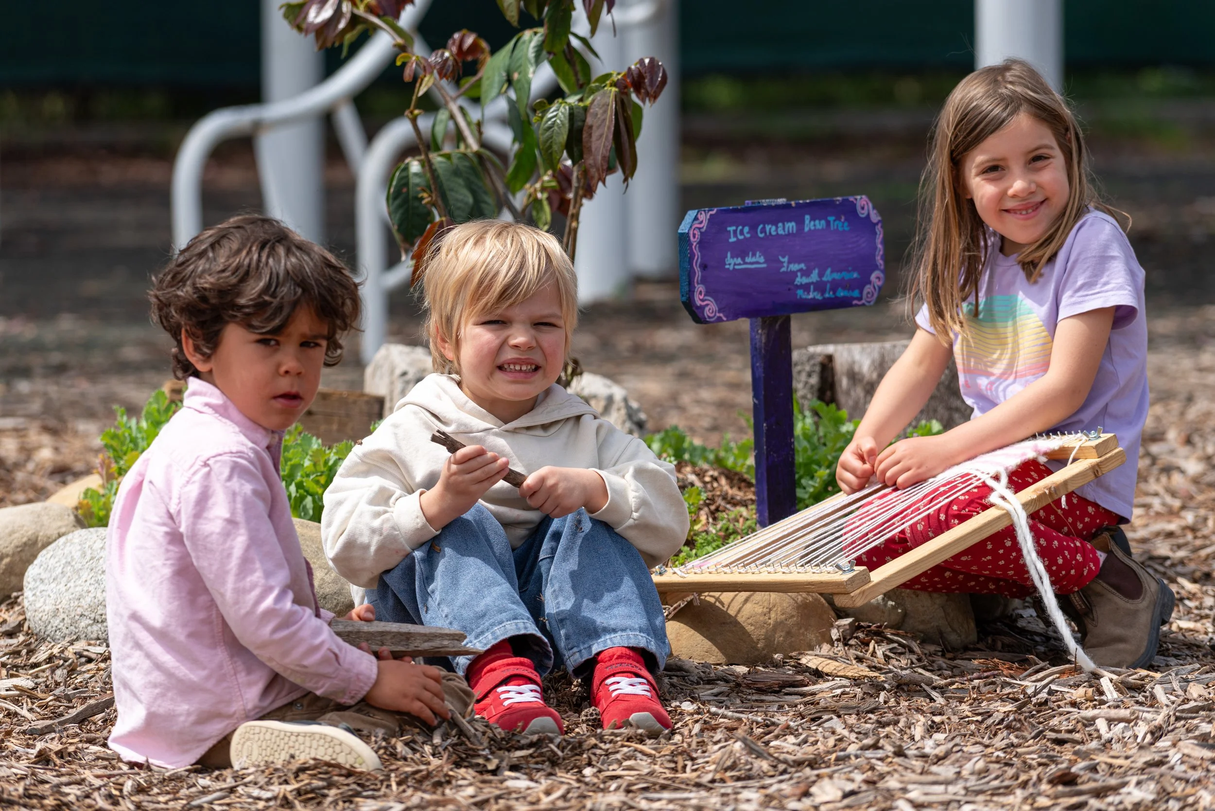 Three children sitting outdoors on wood chips, with a small garden and a purple sign behind them. One girl is sitting on the right, smiling, holding a loom. Two boys are sitting on the left, one looking at the camera with a curious expression, the ot