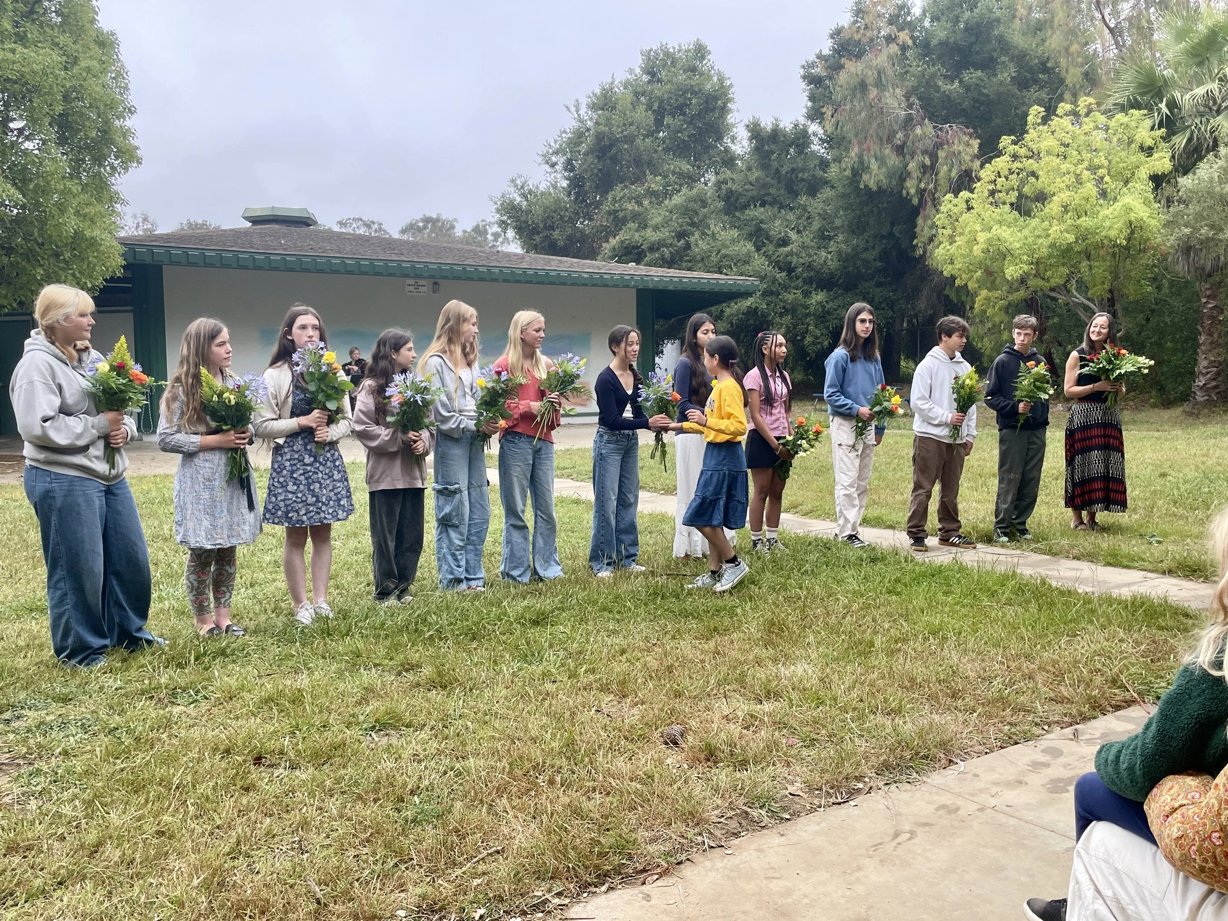 A group of 14 children and one adult woman standing outdoors in a line, each holding a bouquet of flowers, during a school or community event in a park or garden area.