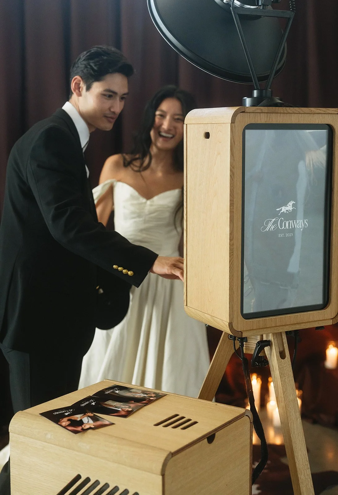A man and woman in formal attire are using a photo booth at a wedding reception. The booth has a wooden frame with a screen displaying 'The Conways, EST. 2025,' along with a dog logo. The woman in the background is smiling and wearing a white dress, while the man in front is touching the booth's screen.