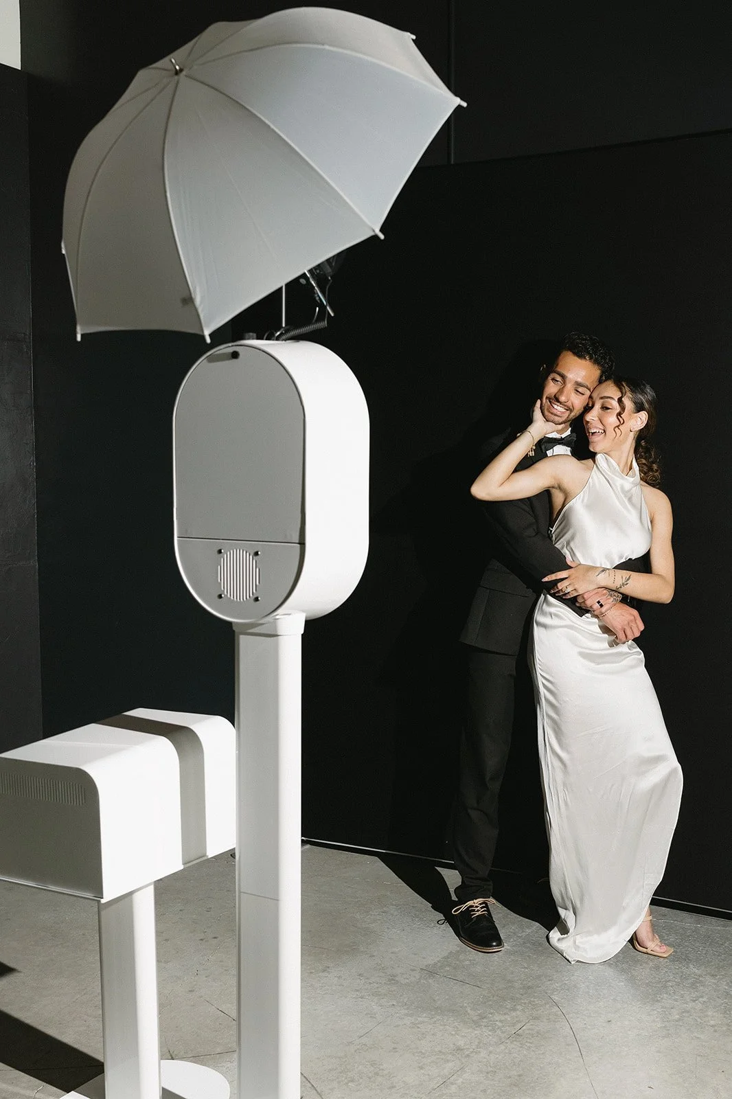 A man and woman dressed in formal attire posing playfully in a photo booth with a black background and a white umbrella-shaped light above them.