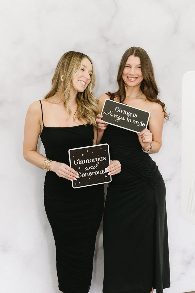 Two women in black dresses holding signs that read 'Glamorous and Generous' and 'Giving is always in style', smiling against a light-colored wall.