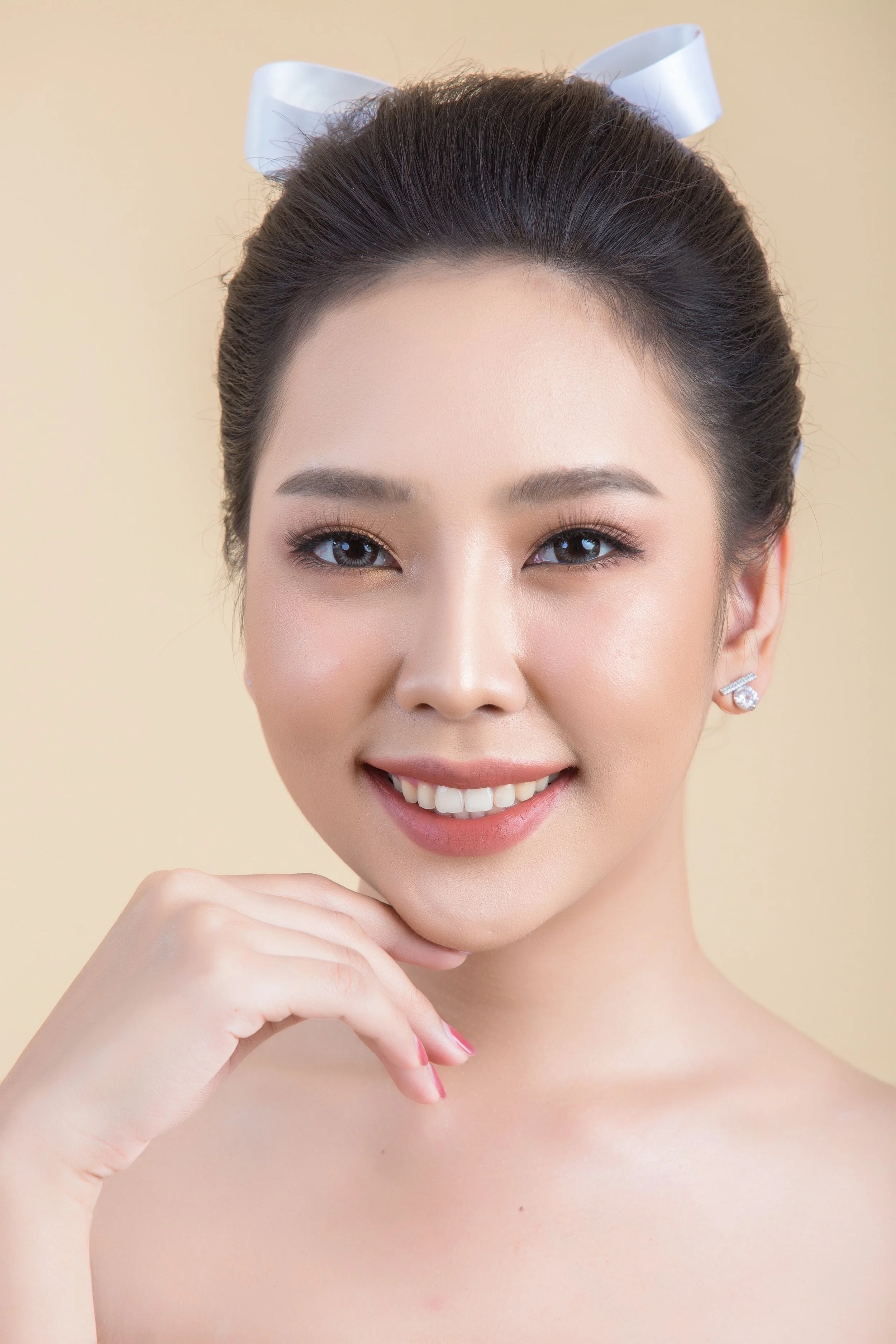 A woman with dark hair styled in an updo, wearing a white bow, smiling with her hand resting on her chin, wearing earrings, makeup, and no visible clothing.