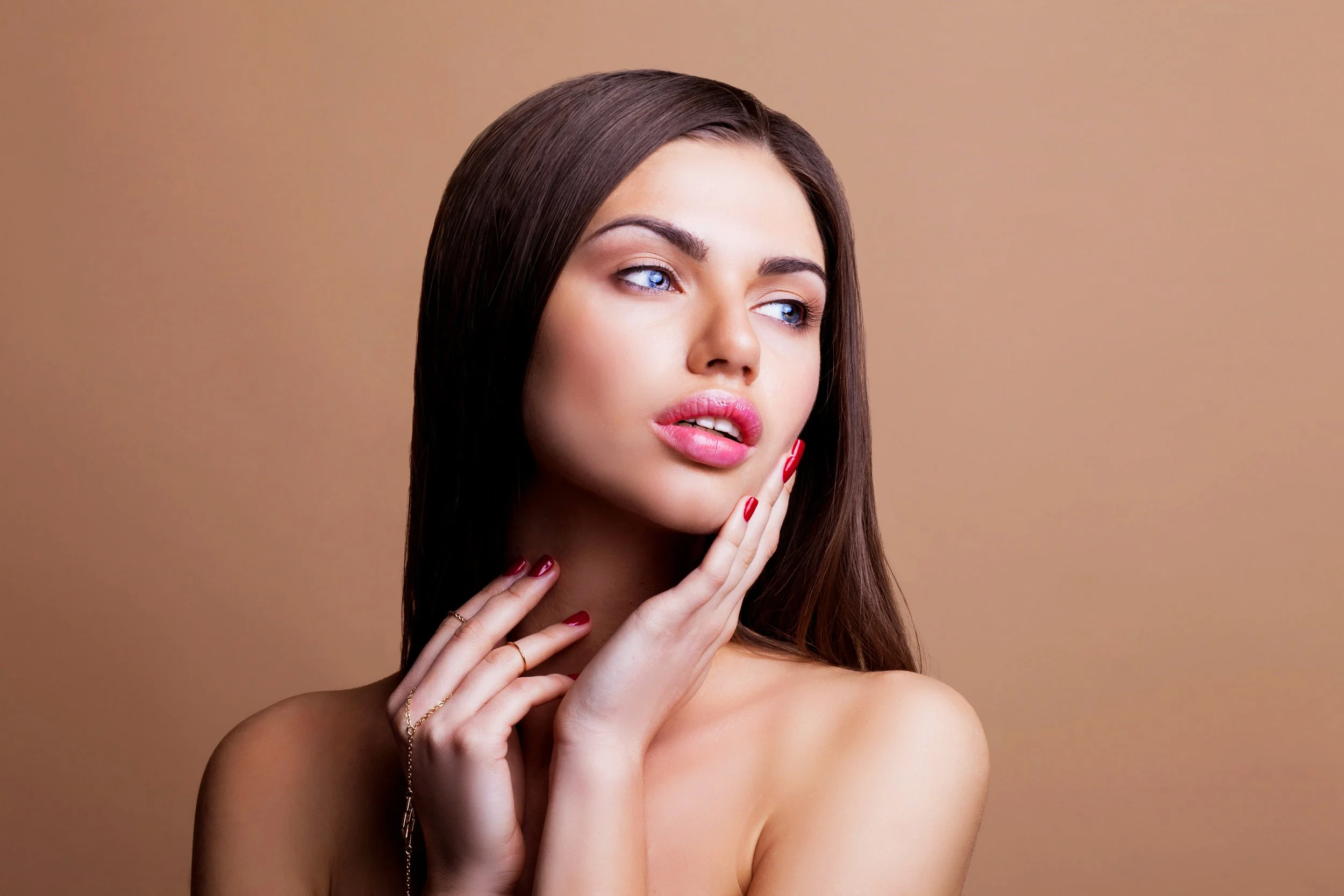 Portrait of a young woman with long brown hair, blue eyes, touching her face with red nail polish, against a plain beige background.