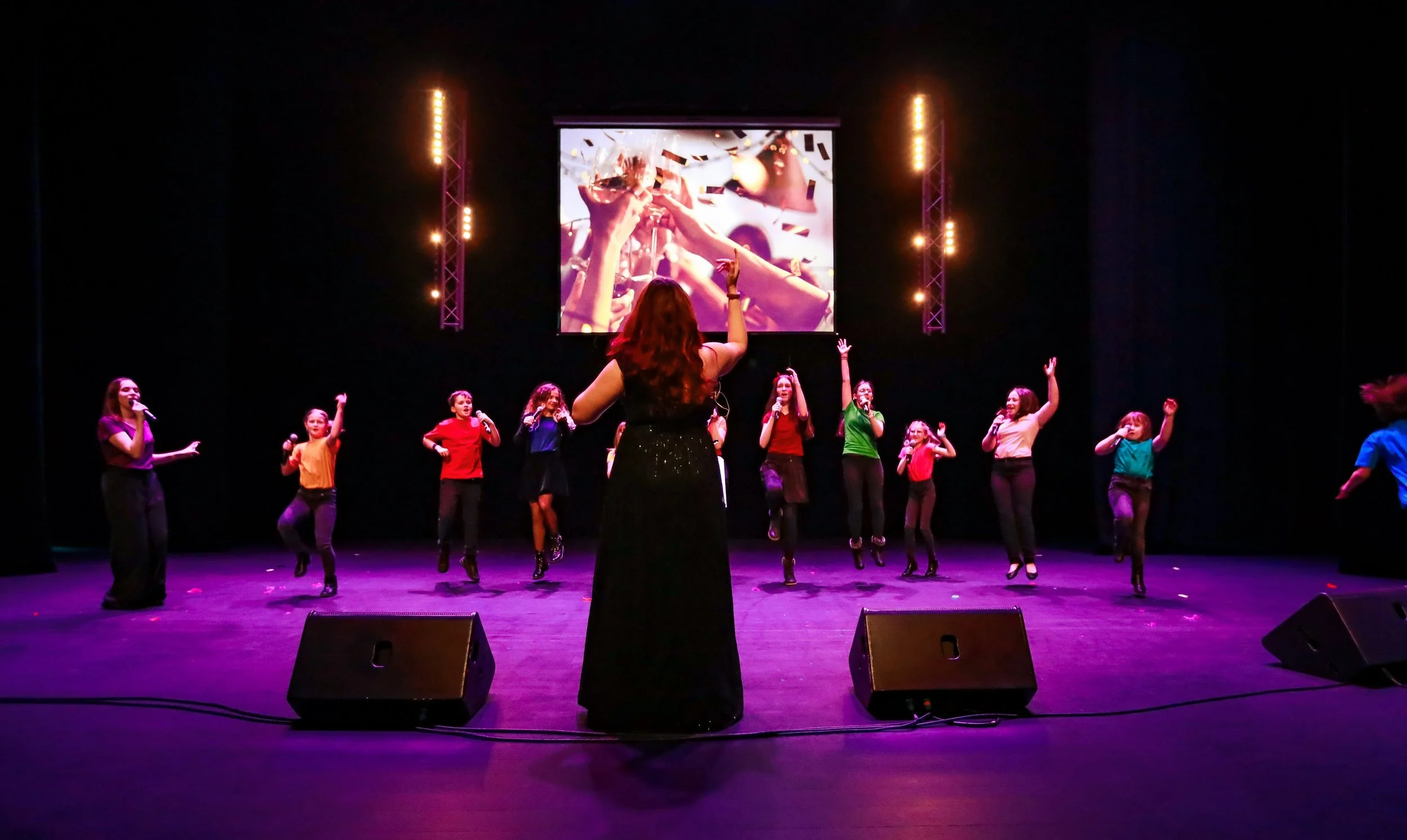 Groupe de jeunes filles en costume coloré chantant et dansant sur scène avec une femme en robe noire, sous un écran géant montrant des confettis, scène éclairée par des lumières