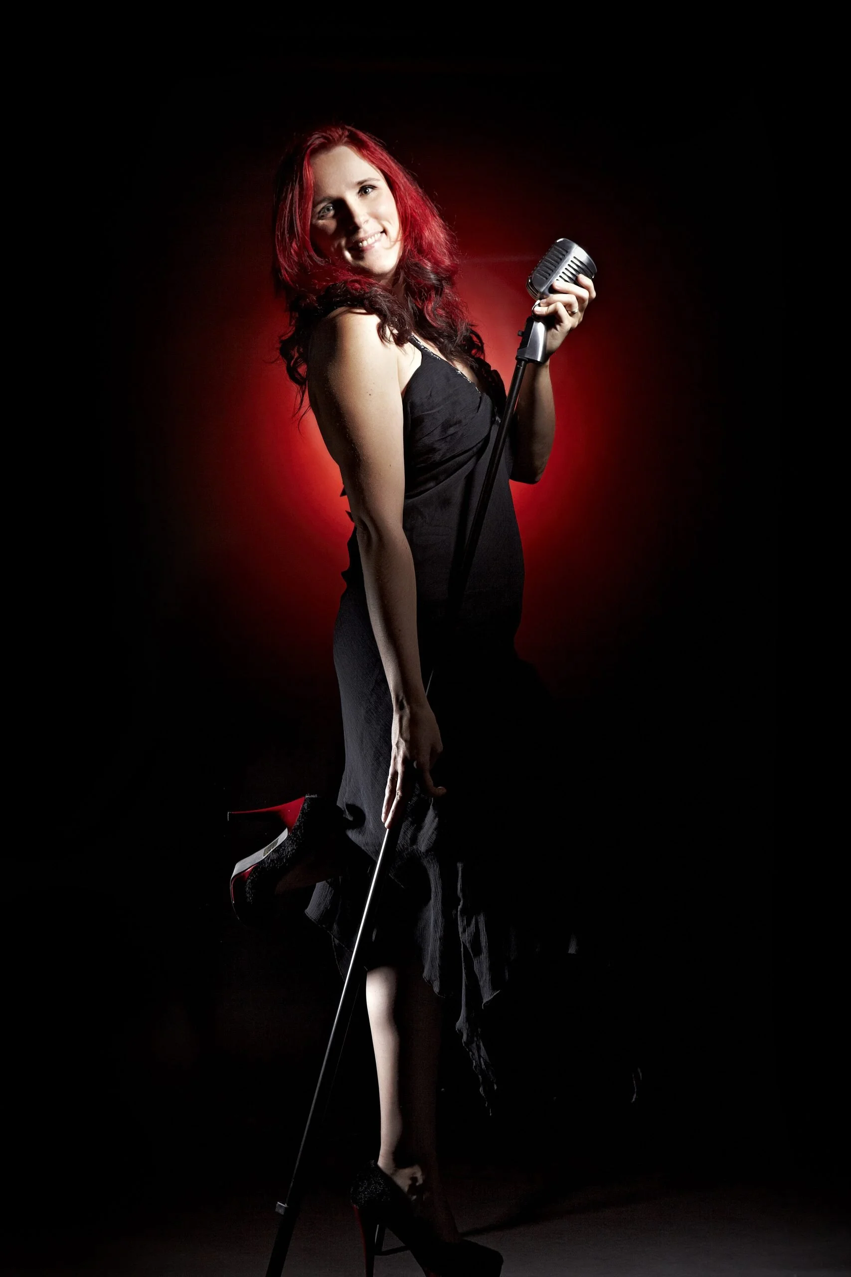 Femme avec cheveux rouges, habillée en noir, posant avec un micro et des talons haut à platforme, fond sombre avec une lumière rouge derrière elle.