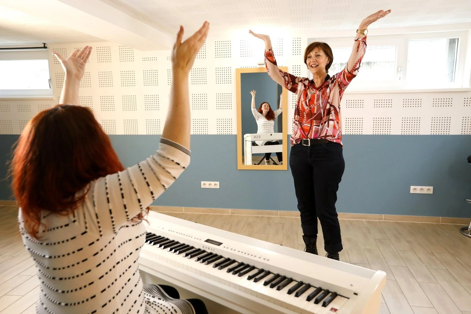 Deux femmes, l'une jouant du piano et l'autre chantant ou gesticulant, dans une salle de musique ou de répétition, avec une grande cloison en arrière-plan.