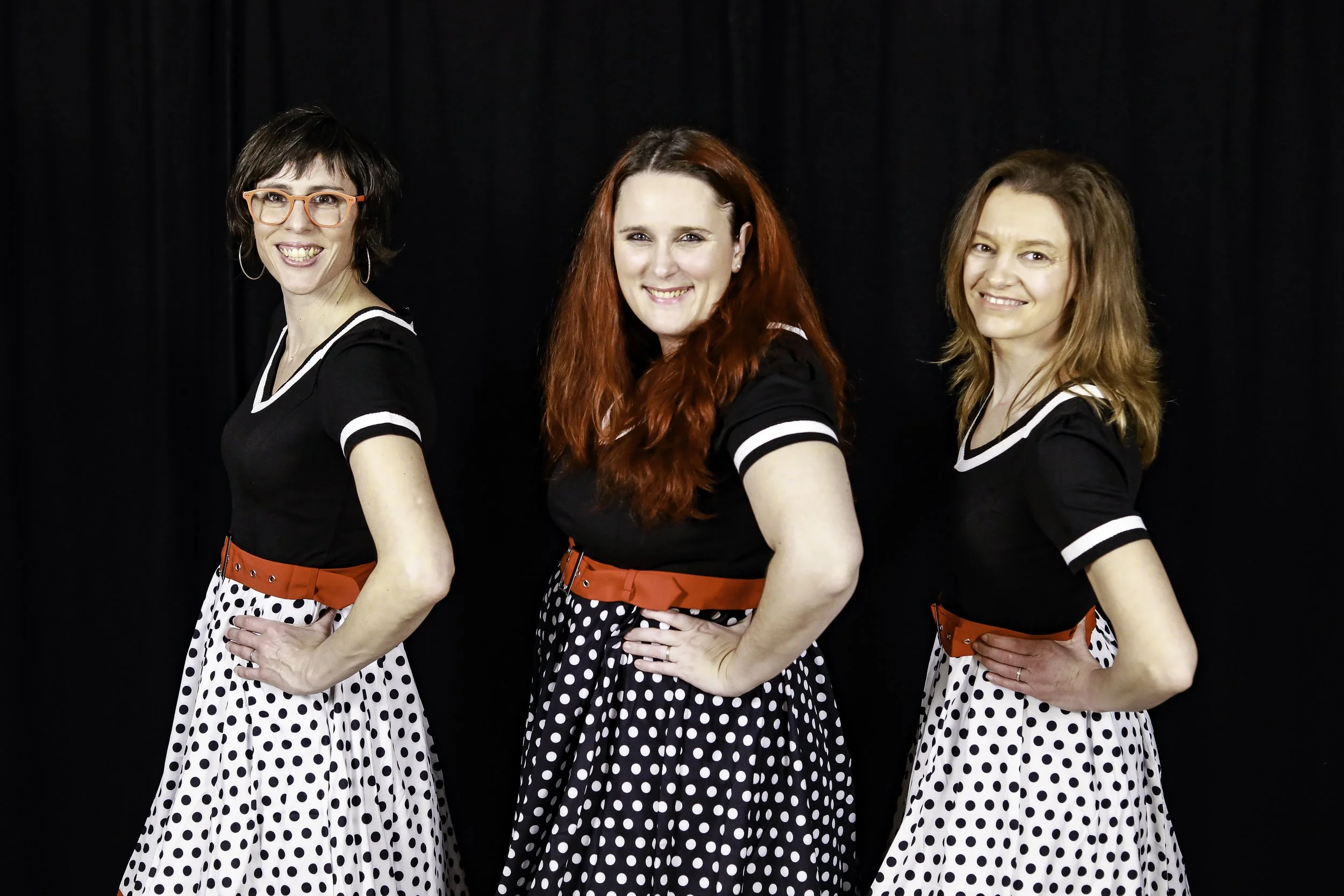 Trois femmes souriantes portant des robes noires avec des jupes à pois noirs et blancs, contre un fond noir.