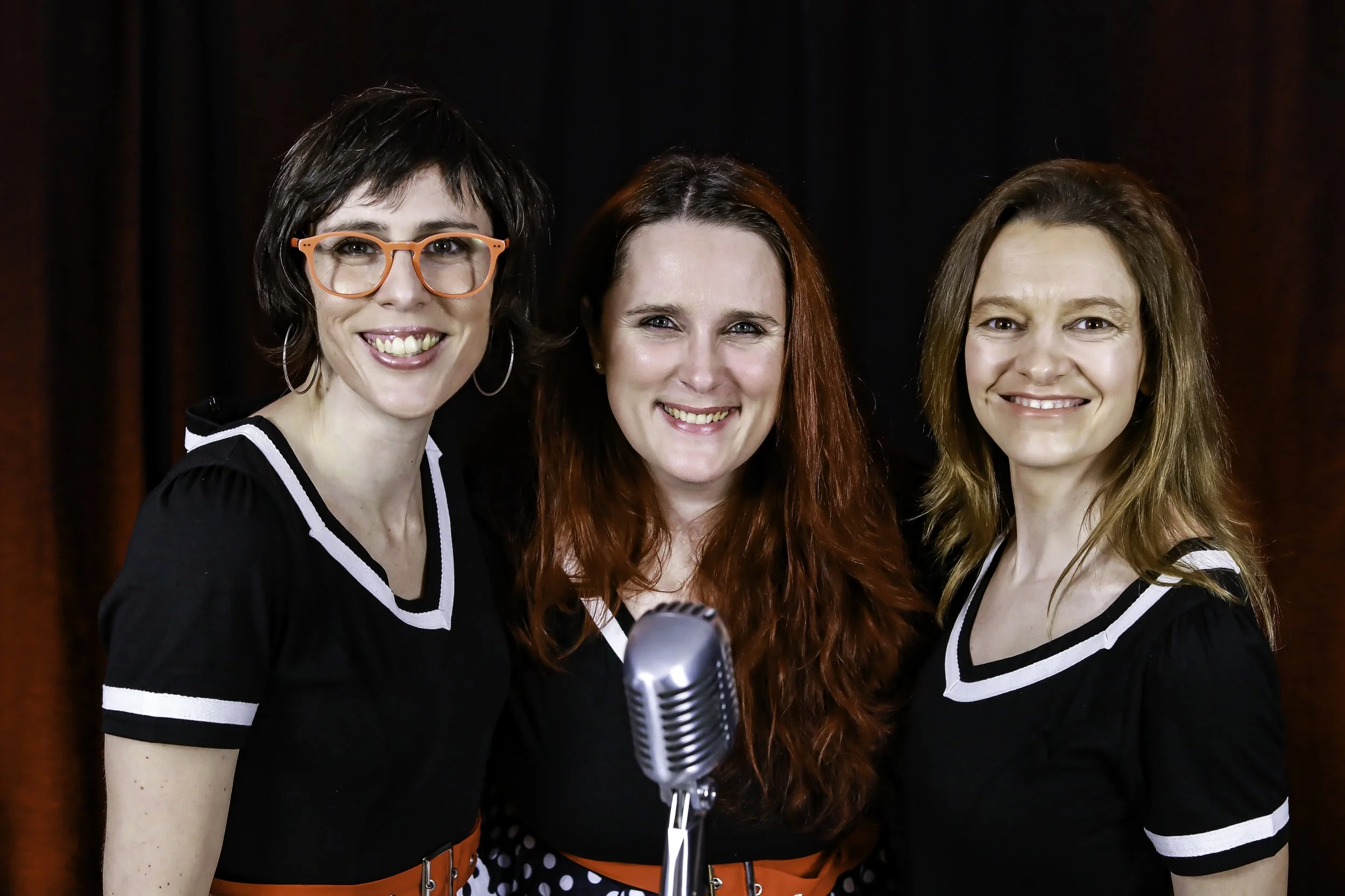 Trois femmes souriantes posent devant un microphone, portant des robes noires avec des détails blancs et rouges, avec un fond noir et rouge.