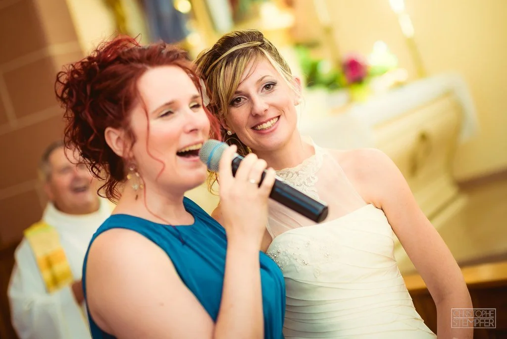 Deux femmes souriantes lors d'une célébration ; l'une chante dans un micro, l'autre porte une robe blanche
