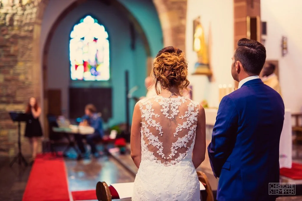 Mariée et marié lors de leur cérémonie dans une église avec un vitrail coloré en arrière-plan.