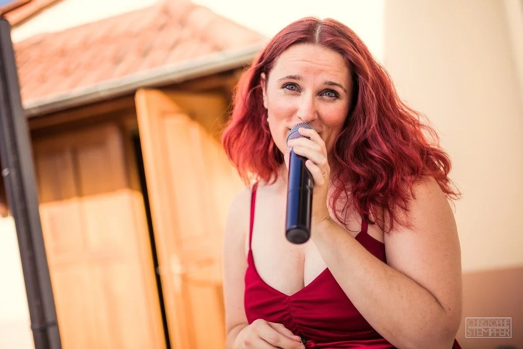 Une femme aux cheveux roux, portant une robe rouge, tient un micro et semble en train de chanter ou de parler.