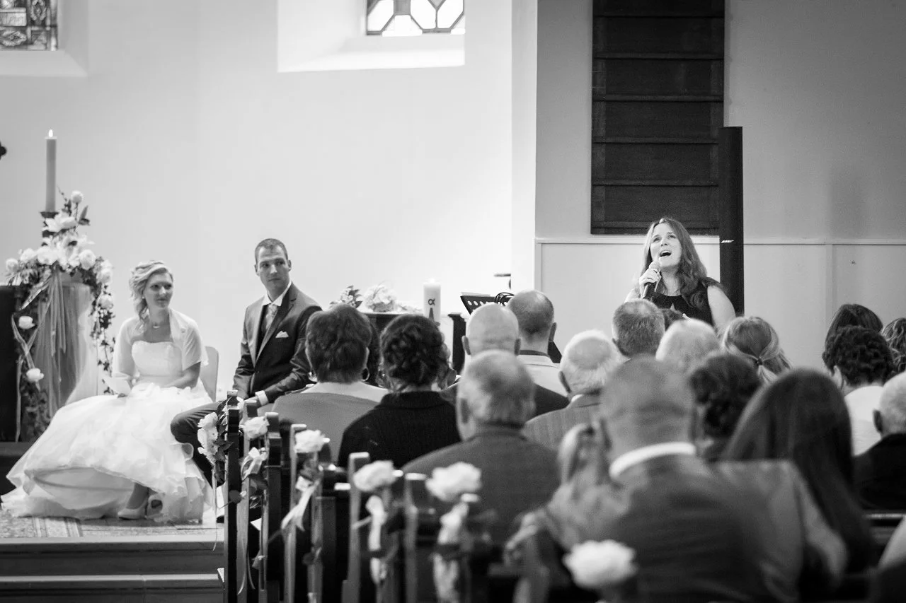Un couple de mariés assis devant des invités lors d'une cérémonie de mariage en intérieur, avec une femme parlant ou chantant à un micro. La scène est en noir et blanc.