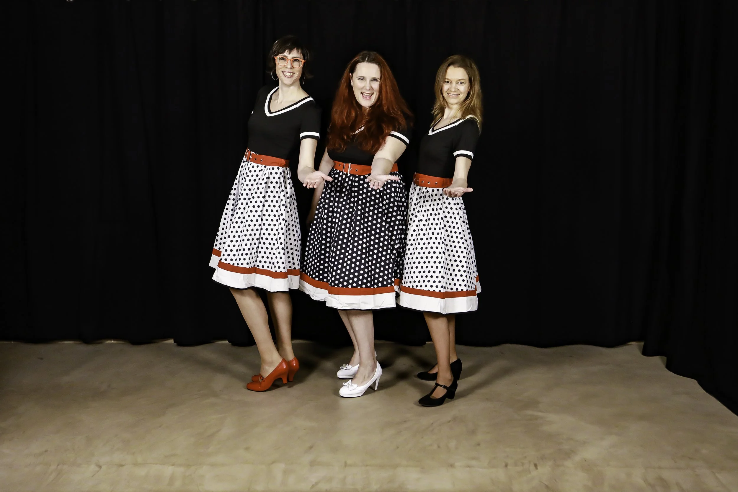 Trois femmes habillées en jupes à pois noirs et blancs avec des hauts noirs, posant devant un rideau noir, souriantes et gesticulant.