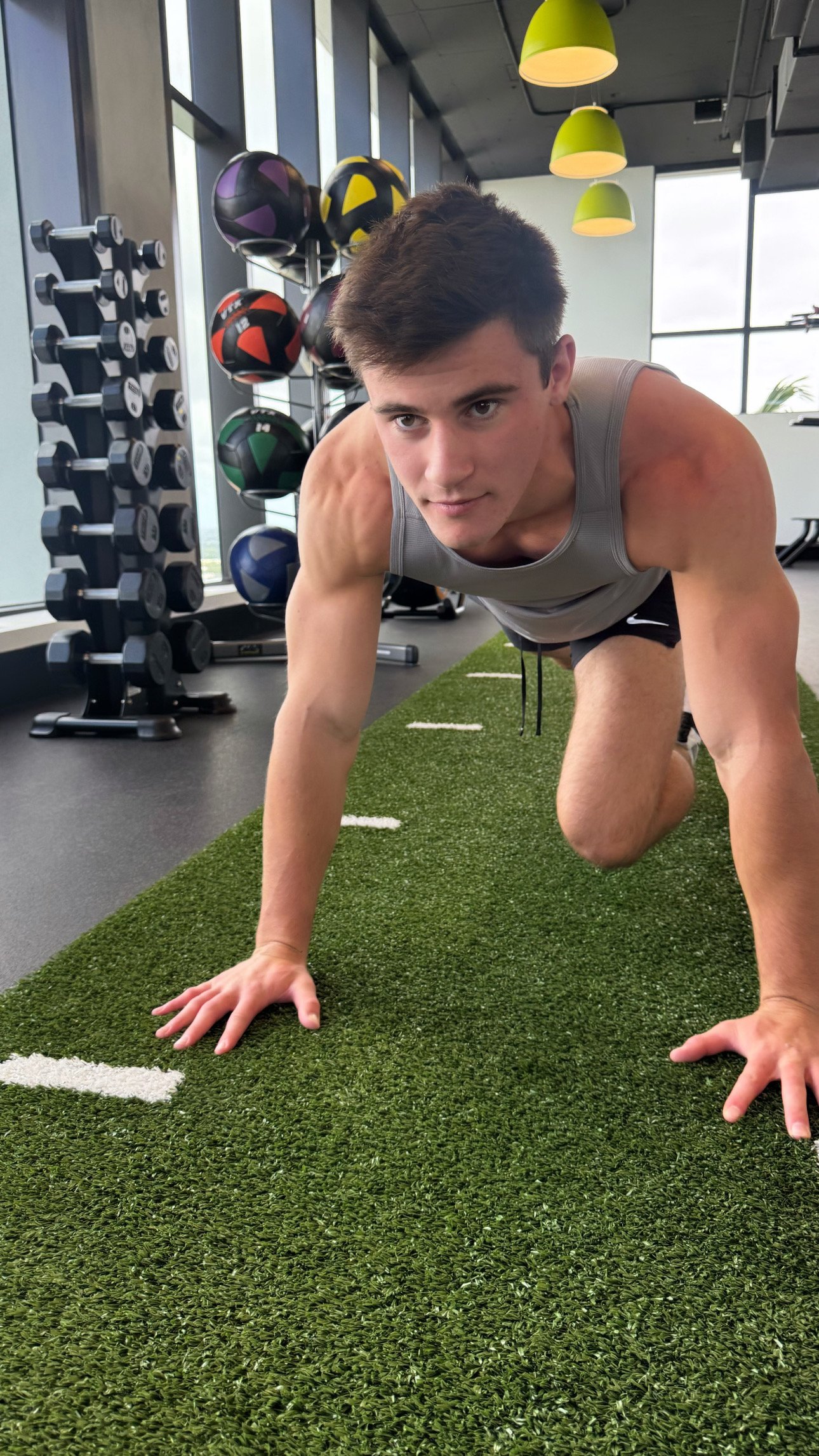 A physically fit man in workout attire on a green turf mat at a gym, in a crawling position, with exercise balls and dumbbells in the background.