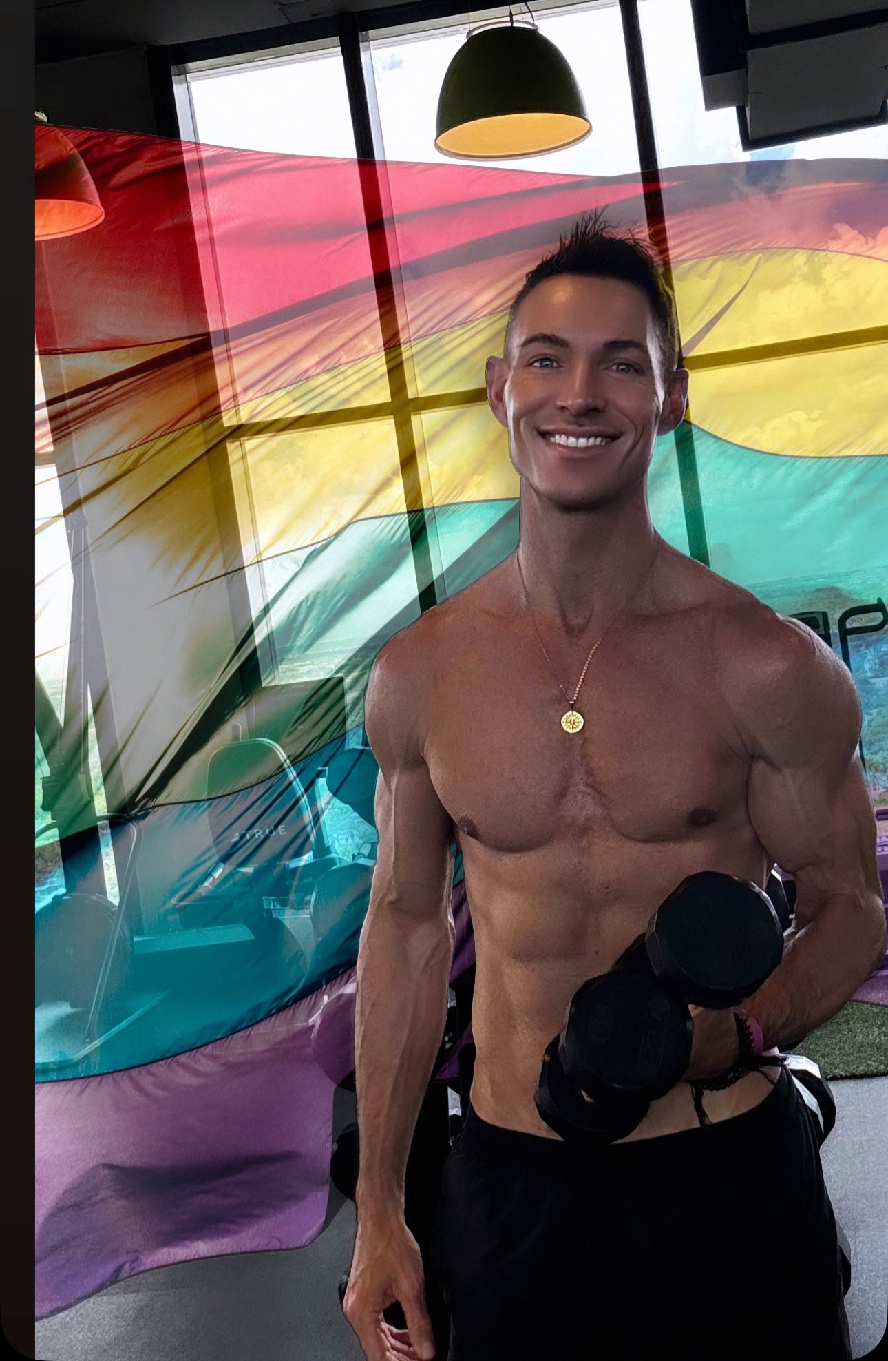 A smiling shirtless man holding a dumbbell in a gym with rainbow-colored rainbow flag in the background.