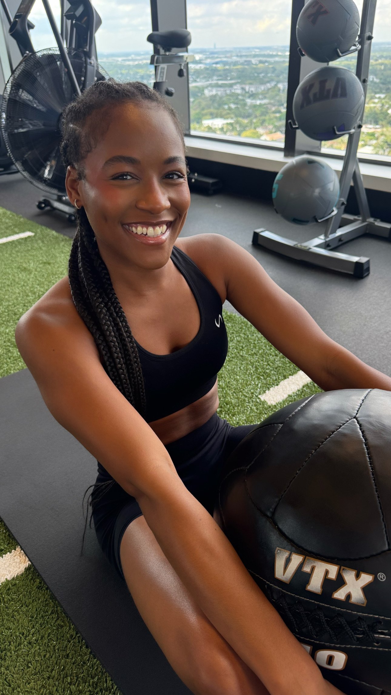 A young woman with braided hair smiling while sitting on a black workout mat in a gym, with a medicine ball labeled VTEX in front of her, gym equipment including medicine balls and a fan in the background, and large windows showing a cityscape view.