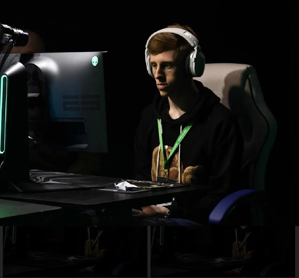 Young man with headphones sitting at a computer desk in a dark room, focused on the screen, with a gaming monitor displaying a glowing alien head logo.