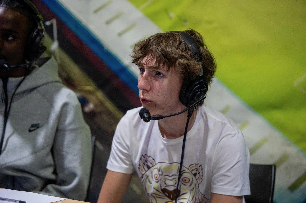 Young man with brown hair wearing a white T-shirt with a tiger face graphic, sitting at a table with a headset on, focused on something in front of him. Part of another person with a gray hoodie wearing a headset is visible next to him.