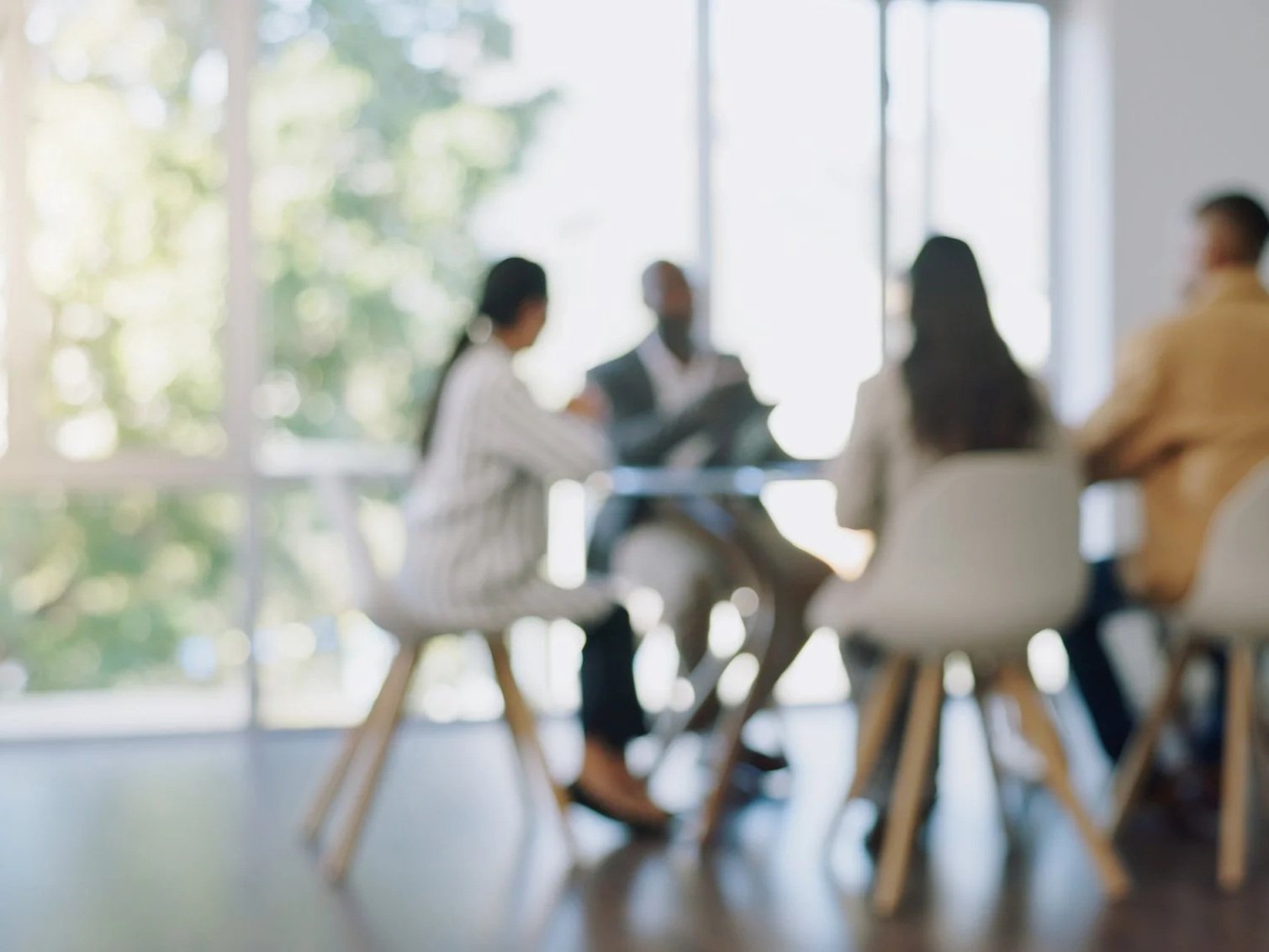 Blurred image of four people sitting around a table in a bright room with large windows, possibly having a discussion or meeting.