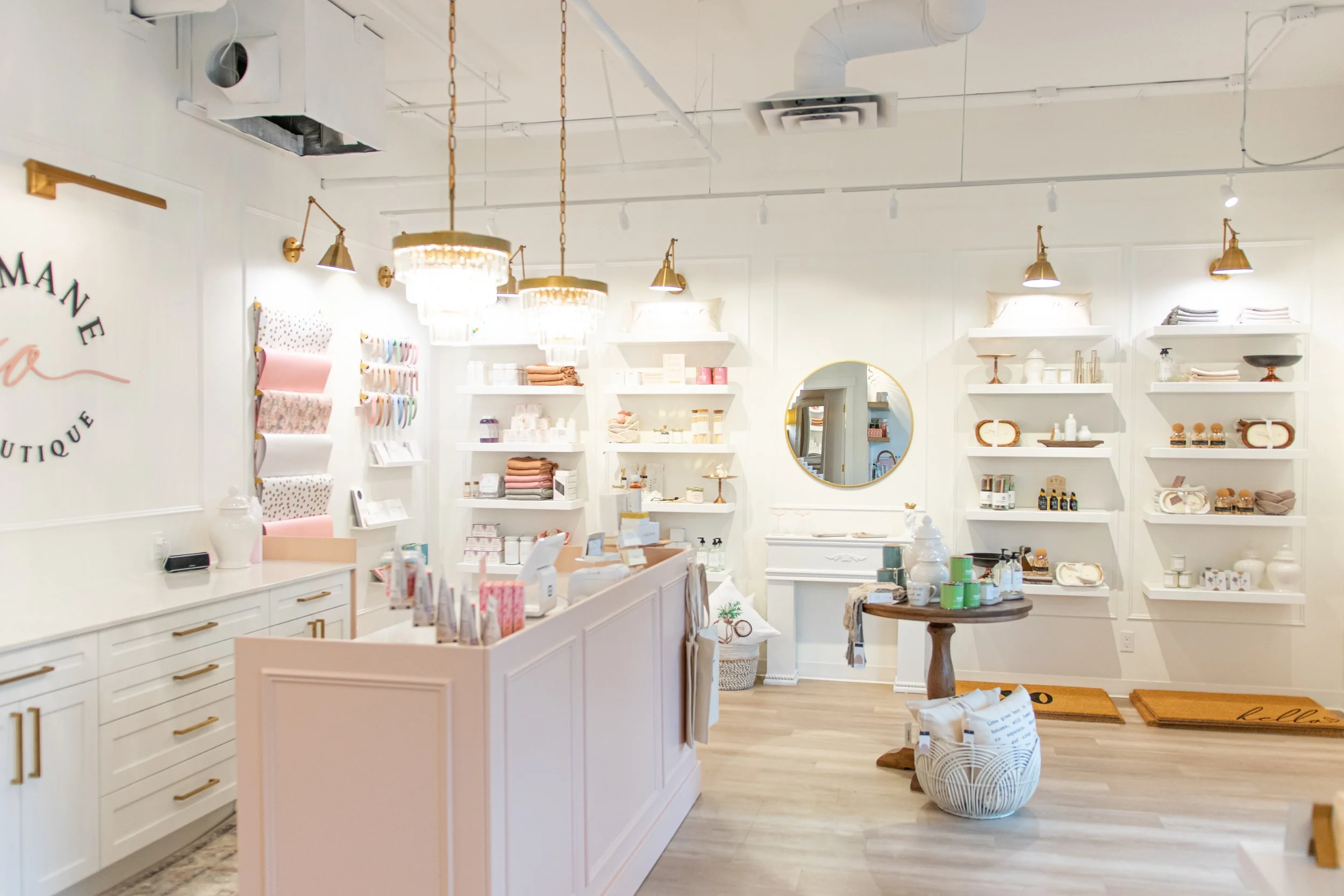 Interior of a boutique shop with white walls, shelves displaying various home decor and gift items, a pink counter, and a round wooden table with products on it. There are chandeliers and spotlights hanging from the ceiling.
