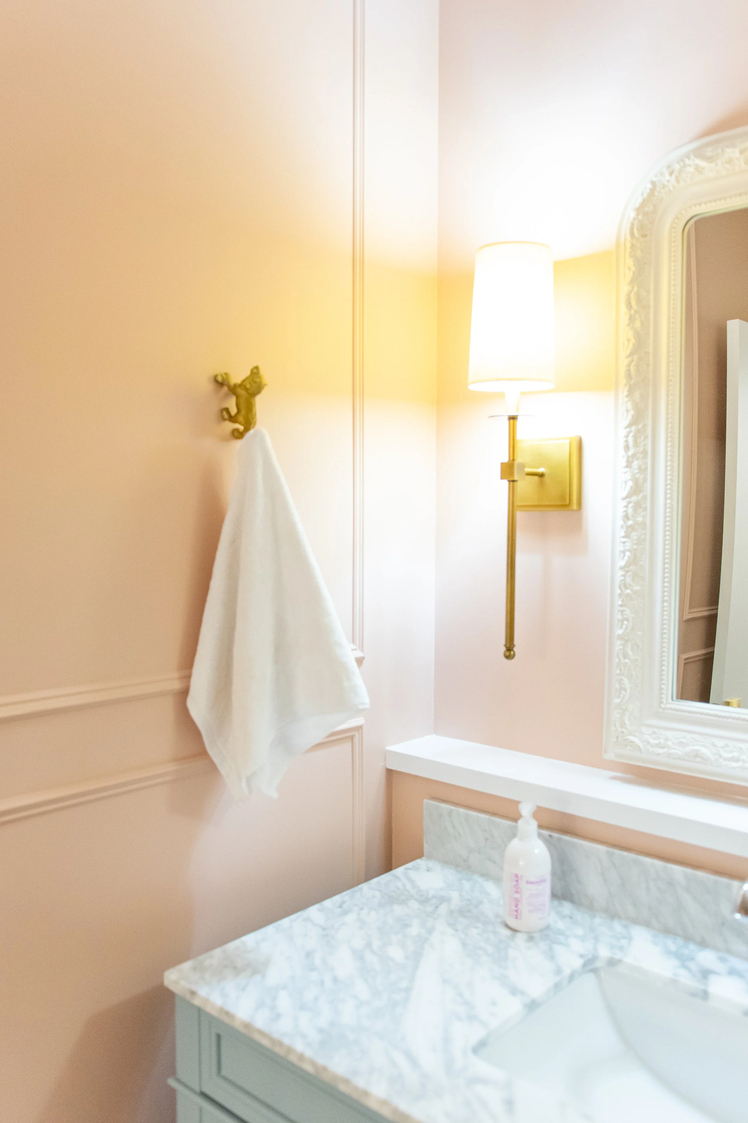 A bathroom with pale pink walls, a white marble countertop, a white towel hanging on a gold hook, a gold wall sconce with a white lampshade, and a white framed mirror.
