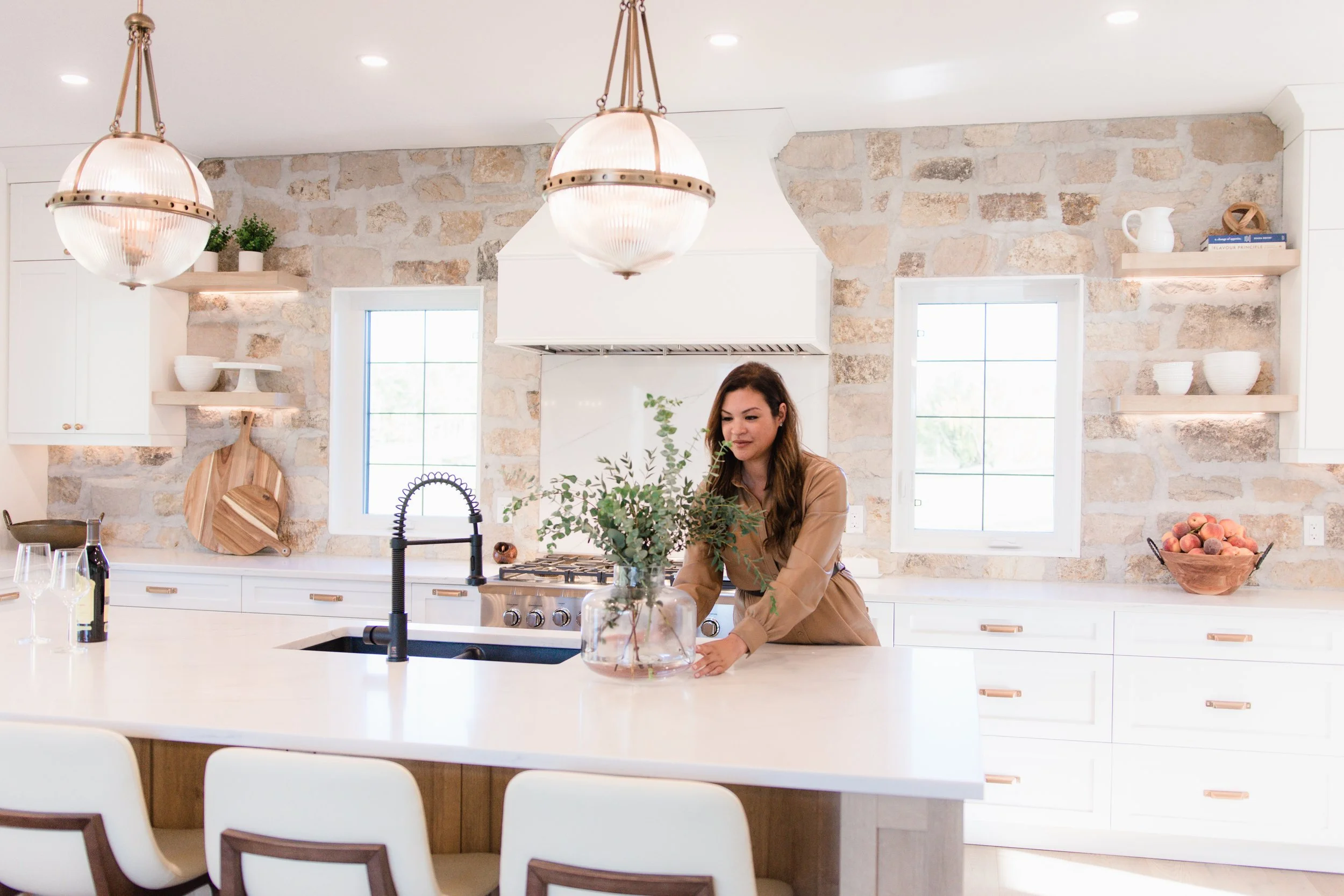 Woman placing a glass vase with green leafy plant on a kitchen island with two white chairs. Kitchen has stone wall, white cabinets, two windows, and pendant lights.