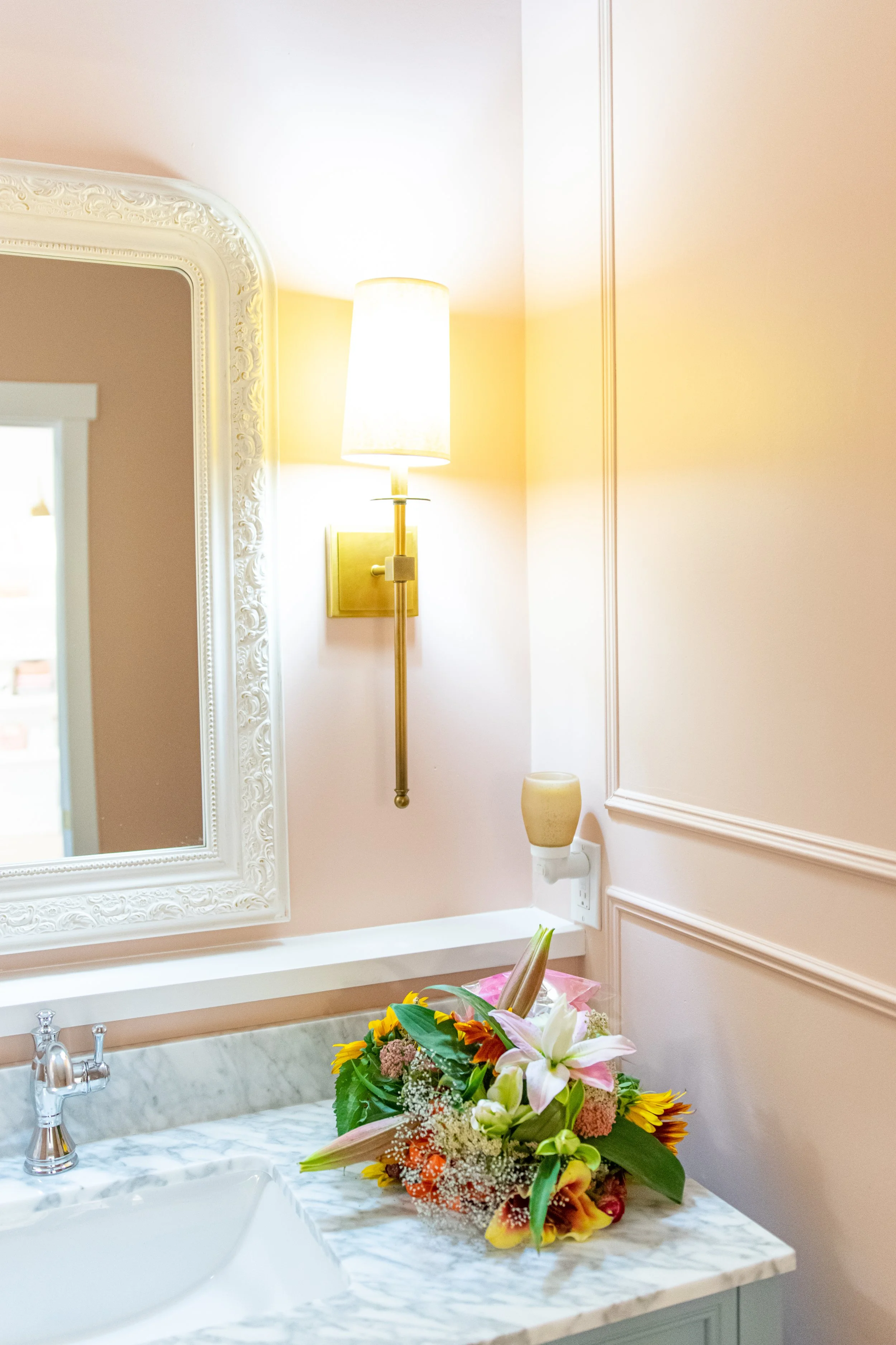A bathroom vanity with a marble countertop holding a colorful flower bouquet, next to a white sink and a silver faucet. A mirror with an ornate white frame is above the vanity. A wall-mounted light fixture with a lampshade is turned on, illuminating 