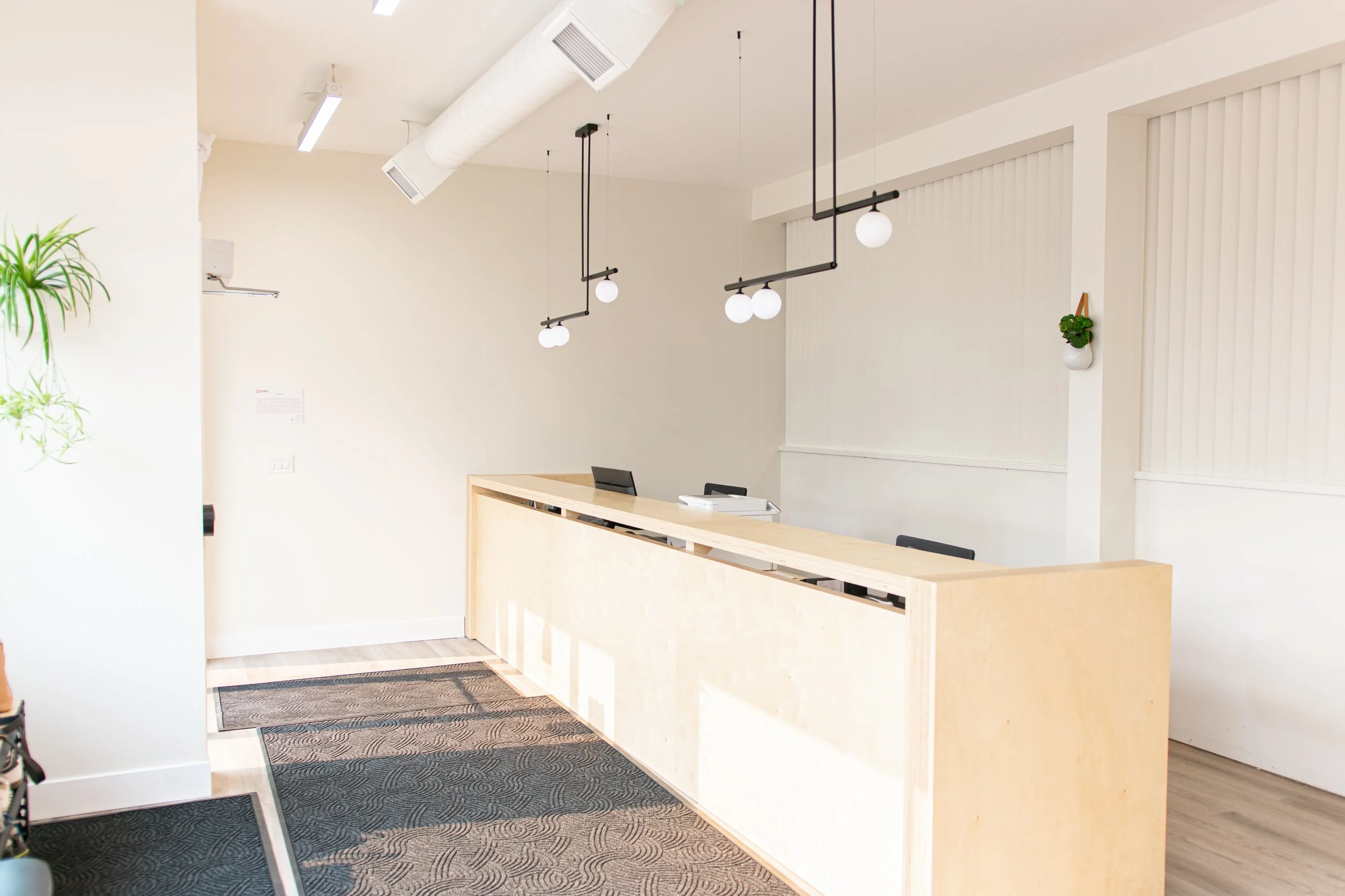 Minimalist reception area with a light wood counter, three black chairs, geometric pendant ceiling lights, and a small potted plant on the wall.
