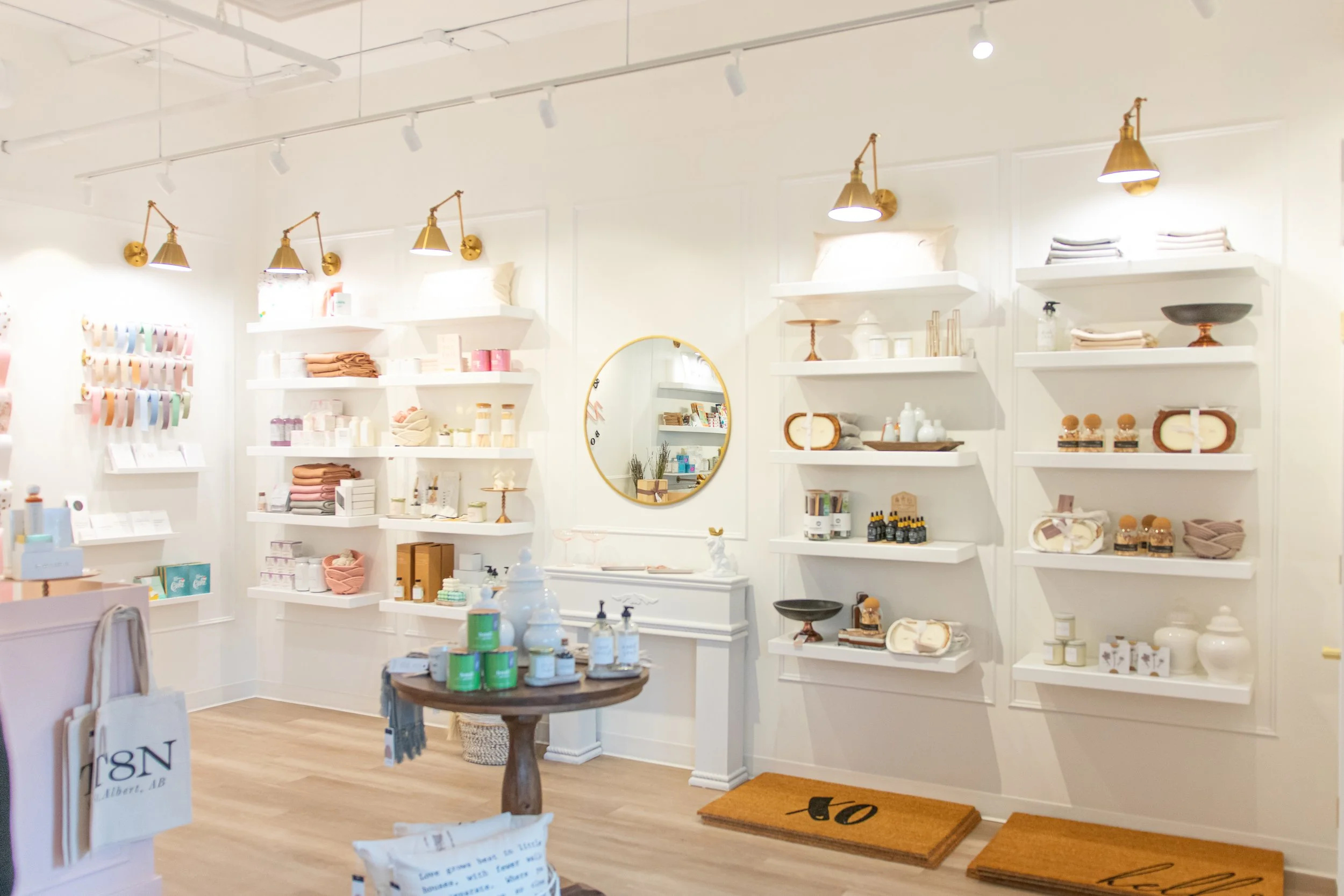 Interior of a modern retail store with white walls and wooden floors, displaying various home decor and gift items on white shelves, including candles, bowls, and textiles, with a round mirror on the wall and a small wooden table with products in the