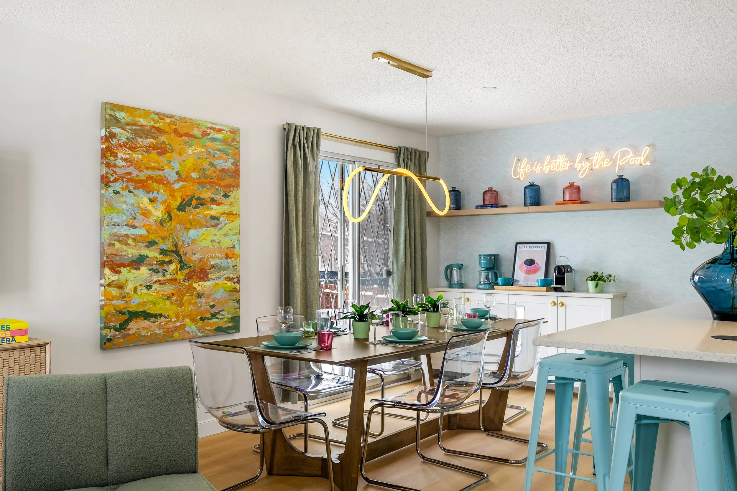 Bright, modern dining area with a wooden table set with turquoise bowls, pink glasses, and small plants. Transparent chairs surround the table. A colorful abstract painting hangs on a white wall, next to a large window with green curtains. The adjacent kitchen features a white counter, blue and pink jars on a shelf, a neon sign saying 'Life is better by the Pool,' and various kitchen appliances and decor.