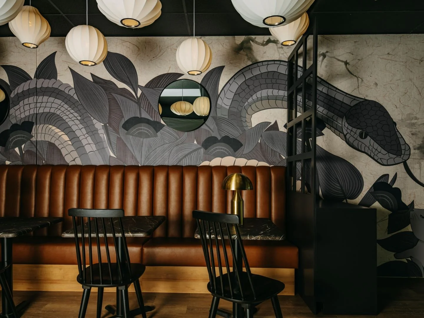 Interior of a stylish restaurant or cafe with black chairs, a brown leather bench, a black marble table with a gold lamp, artistic wall mural featuring a snake and leaves, hanging round white paper lantern lights, and a round mirror on the wall.