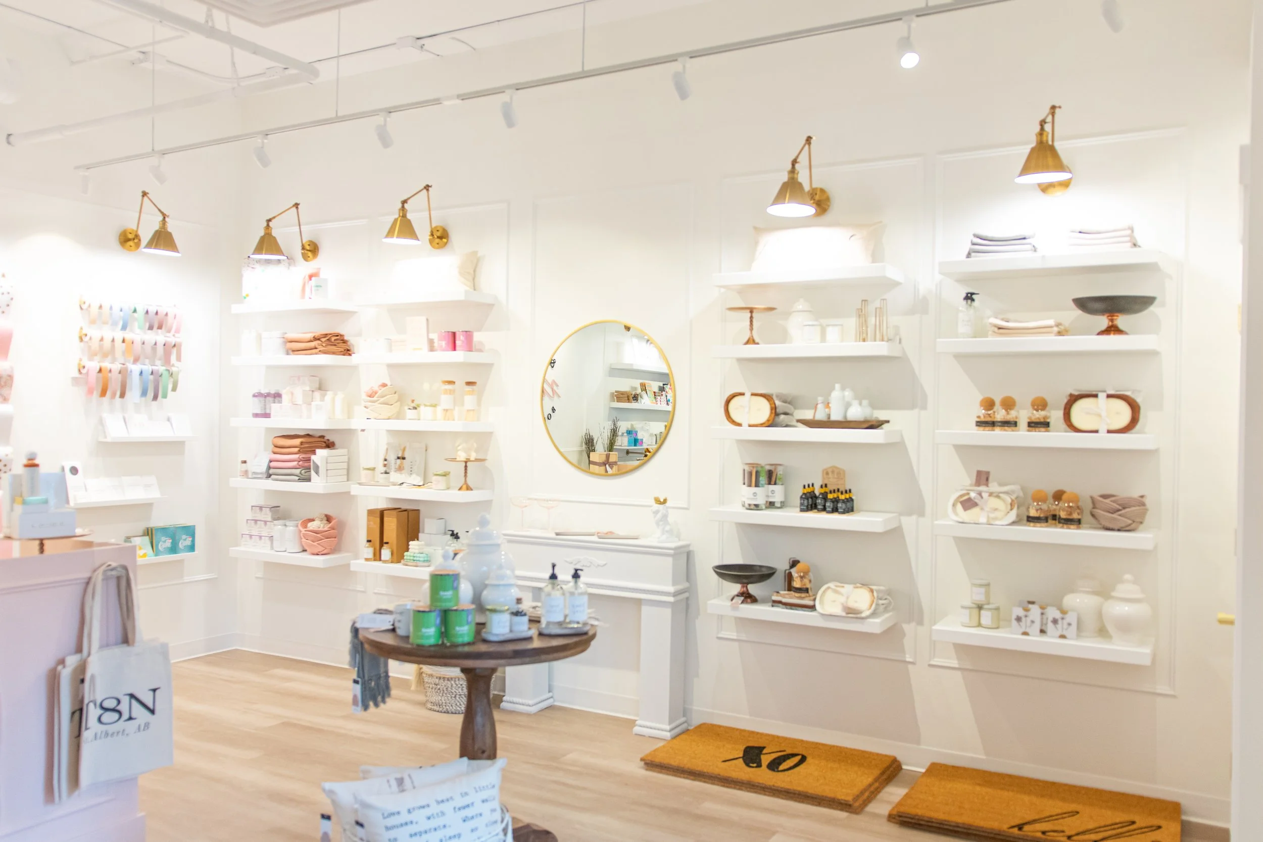 Interior of a retail store with white walls and shelves displaying home decor and accessories, illuminated by brass wall-mounted lights.