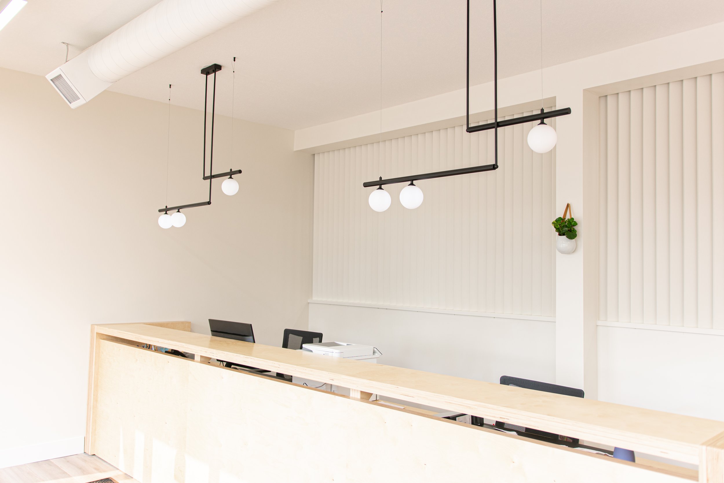 An empty modern office reception area with a light wooden countertop, black chairs, white walls, vertical blinds, pendant lights, an air conditioning vent, and a small potted plant on the wall.