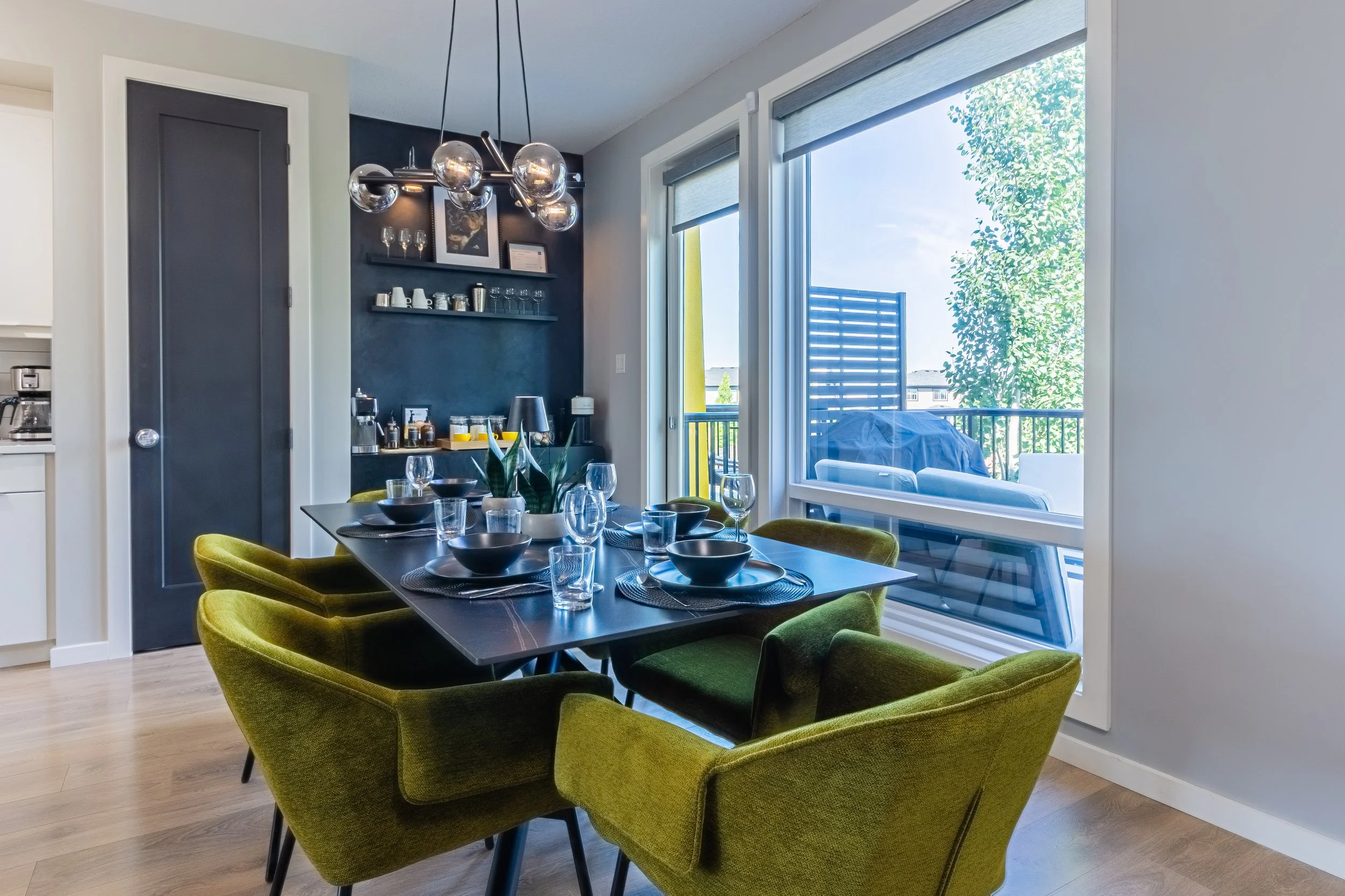A modern dining room with a table set with black bowls, clear glasses, and a centerpiece plant. Surrounding the table are green upholstered chairs. Large windows with blinds look out onto a balcony with outdoor furniture and a barbecue grill, with trees and neighboring buildings visible outside.