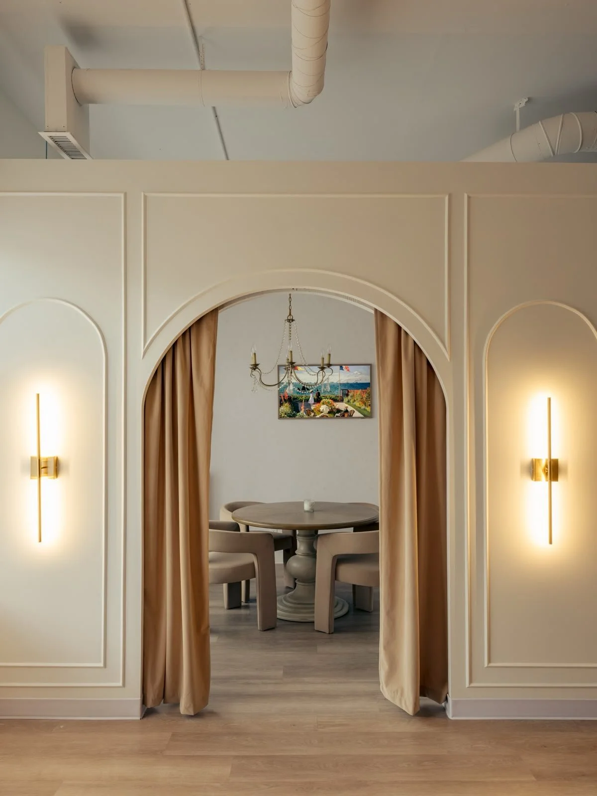 Elegant dining area with a round table, four armchairs, paintings, chandelier, and beige curtains, viewed through an arched doorway with wall sconces.
