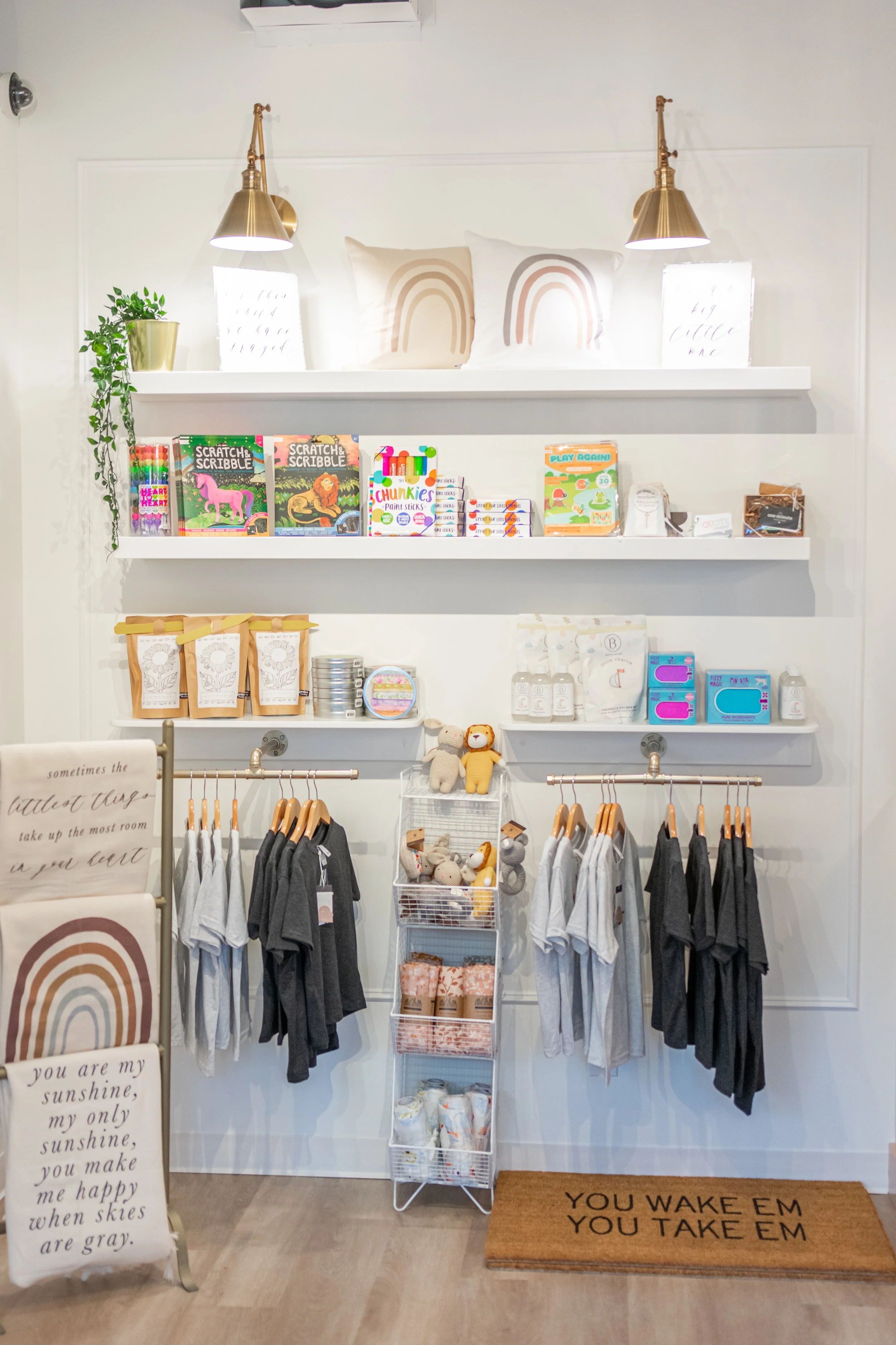 Children's clothing and toys display in a minimalist store with white shelves, hanging shirts, plush toys, and motivational signs on a white wall.