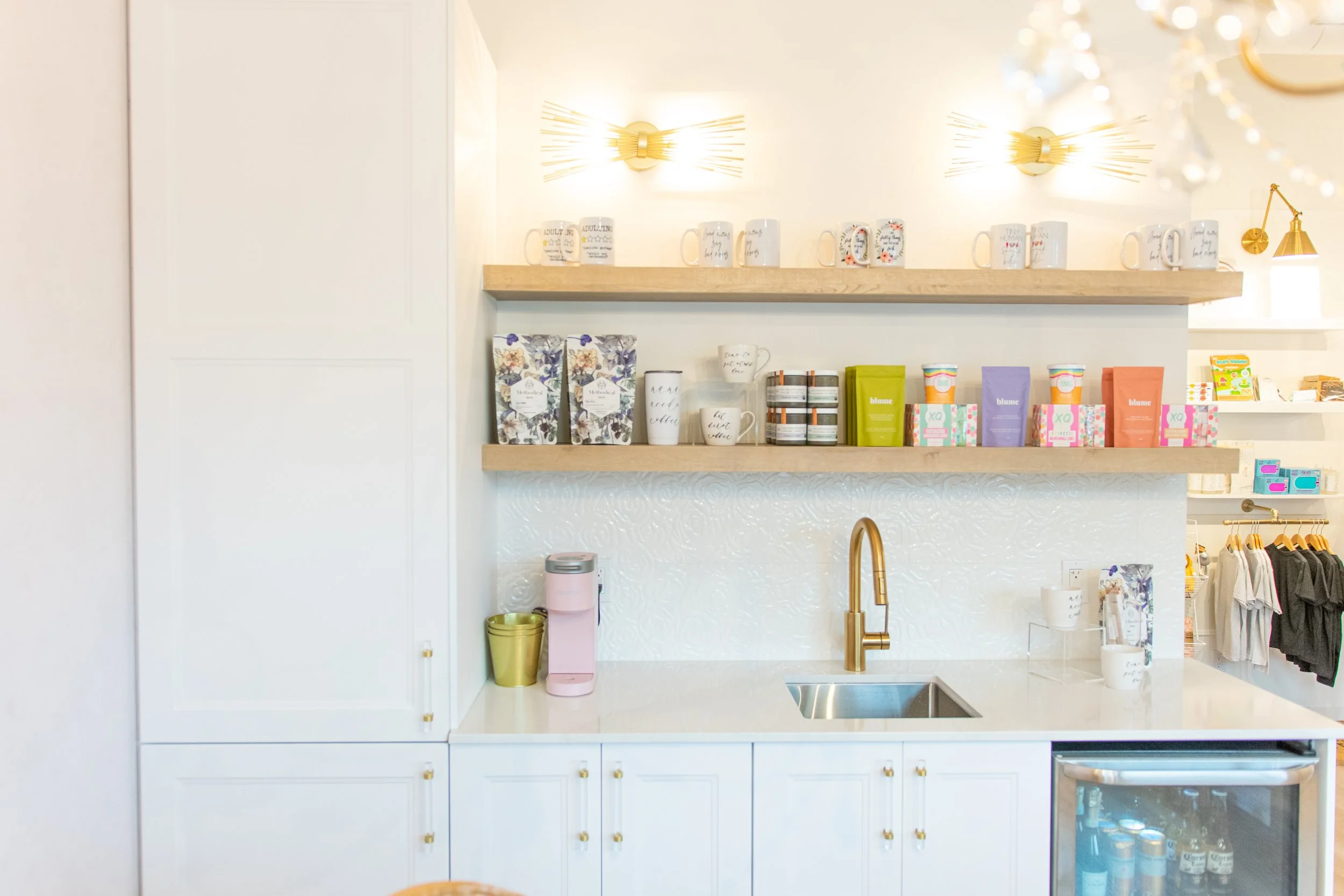 A white kitchen with a gold faucet, white cabinets, and open wooden shelves holding mugs, boxes, and jars. A pink water dispenser and gold trash can are on the counter, with a small sink and a silver refrigerator below.